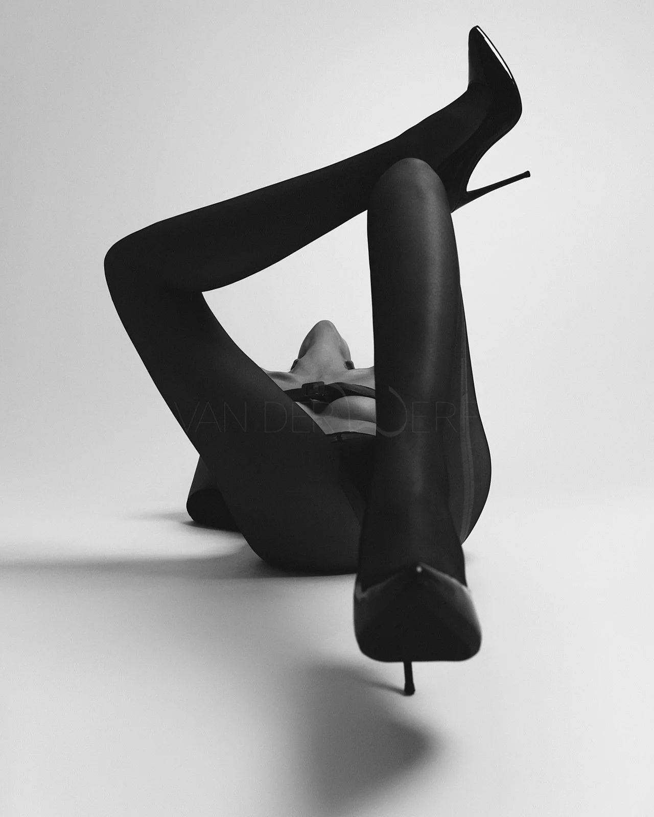 Schwarze hochhackige Schuhe, eine Frau in schwarzem Outfit liegt auf dem Rücken, hebt ein Bein an und hält es hoch.