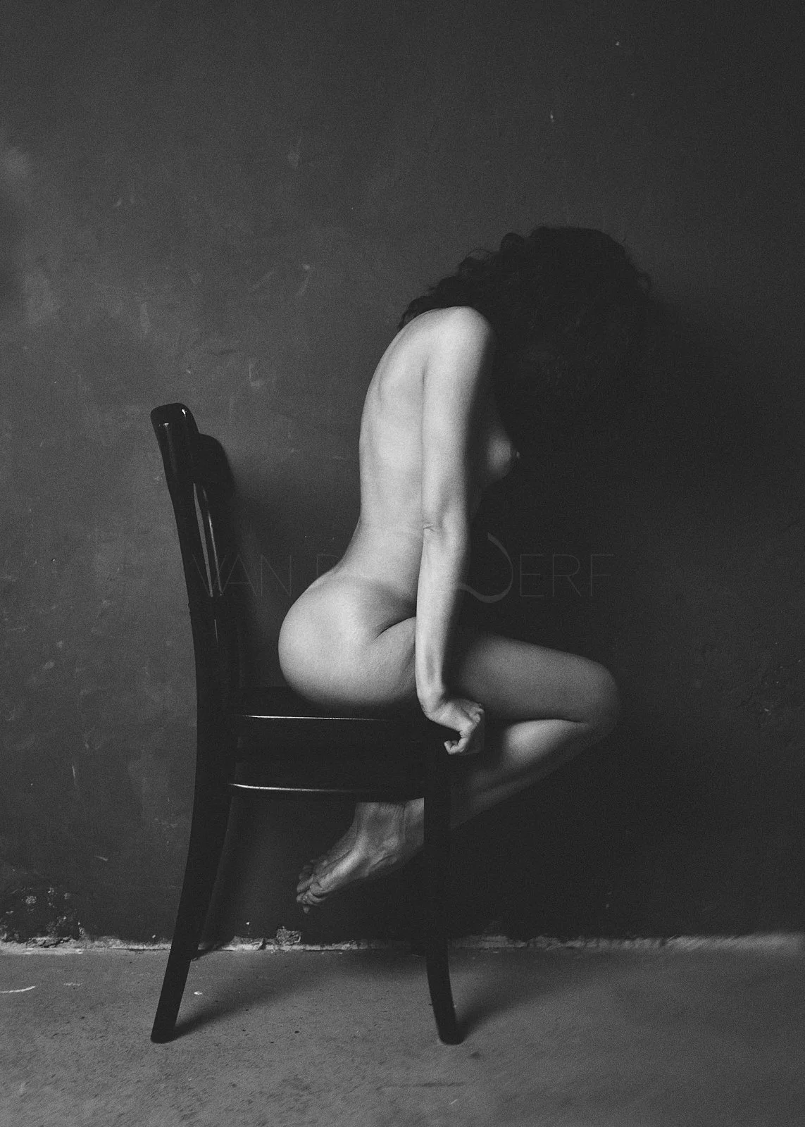 Schwarz-Weiß-Foto eines nackten Menschen mit langen, dunklen Haaren, der auf einem Stuhl sitzt, mit dem Kopf nach unten gebeugt, vor einer dunklen Wand.