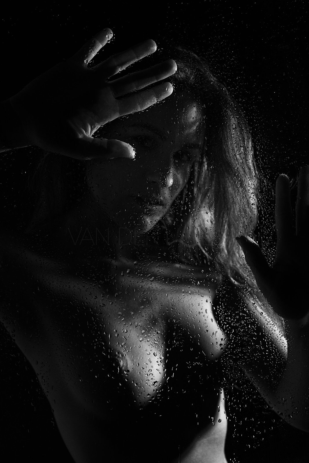 Schwarz-weiß-Fotografie einer Frau, die hinter einem nassen Glas steht. Wassertröpfchen sind auf der Glasoberfläche sichtbar, während die Frau zum Teil durch das Glas sichtbar ist. Sie hebt eine Hand, um das Glas zu berühren oder zu stützen.