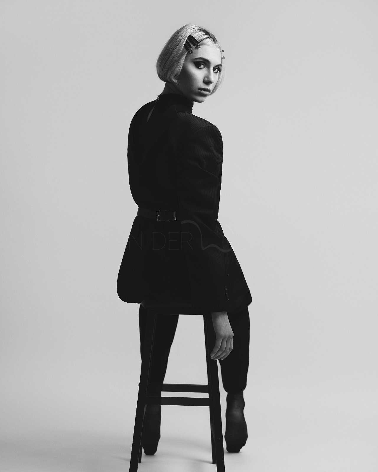 Schwarz-Weiß-Foto eines Models, das auf einem Stuhl sitzt, trägt elegante Kleidung und hat einen Hautansatz mit Haarclips, vor einem hellen Hintergrund.