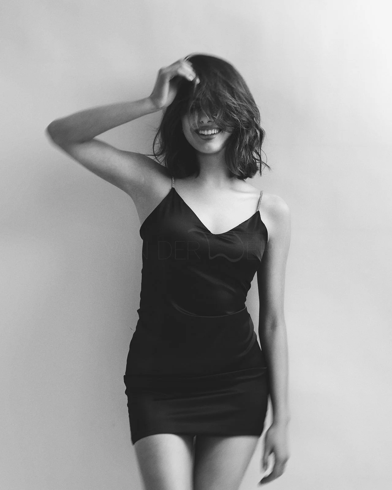 Eine lachende Frau in einem schwarzen, schulterfreien Kleid steht vor einer weißen Wand, mit zerzaustem Haar und einer Hand im Haar.