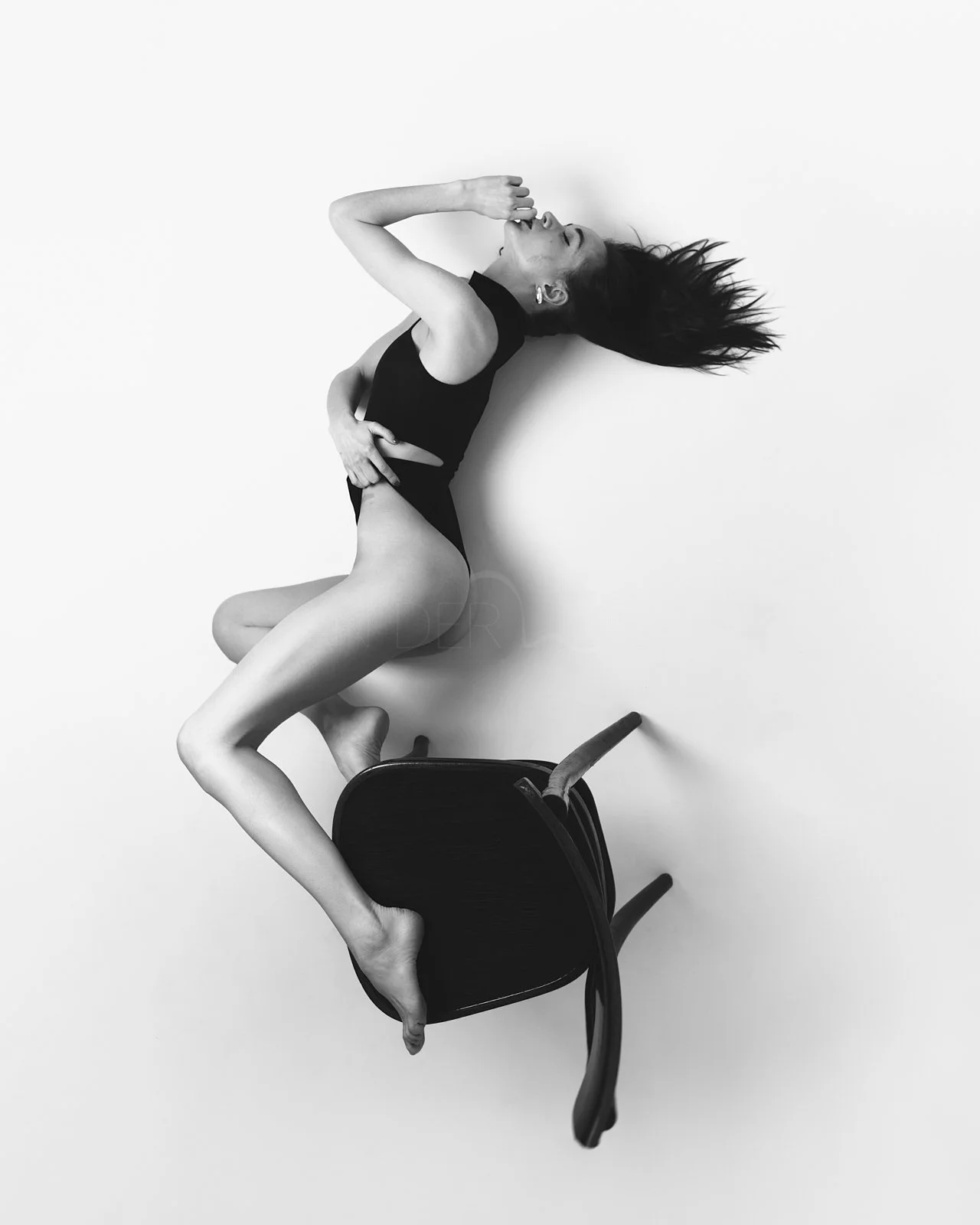 Schwarz-Weiß-Foto einer tanzenden Frau in Schwarzkleidung, die auf einem umgedrehten Stuhl im weißen Raum liegt.