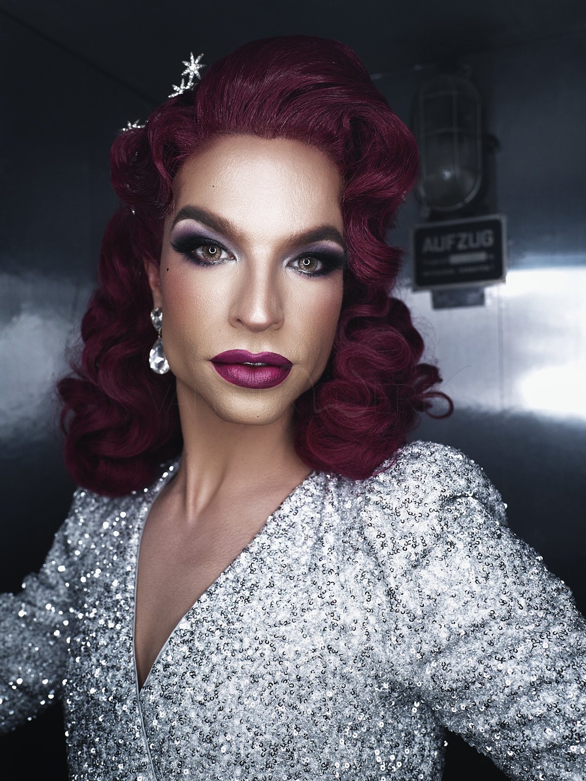 Porträt einer Frau mit dramatischem Make-up, roten Locken, silbernem, glitzerndem Kleid und Ohrringen vor einem dunklen Hintergrund.