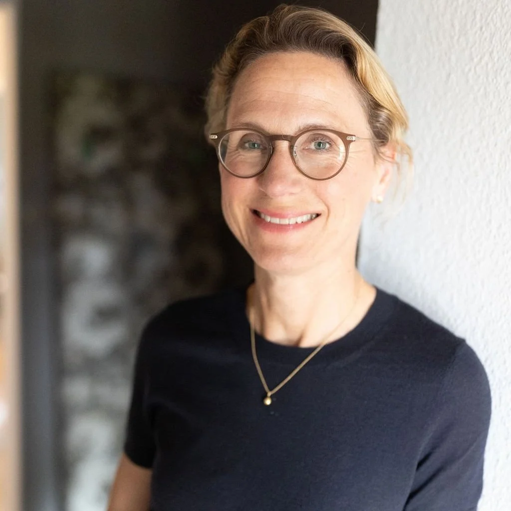 Nahaufnahme einer lächelnden Frau mit Brille, schwarzem Oberteil und goldener Halskette, die vor einer weißen Wand steht.