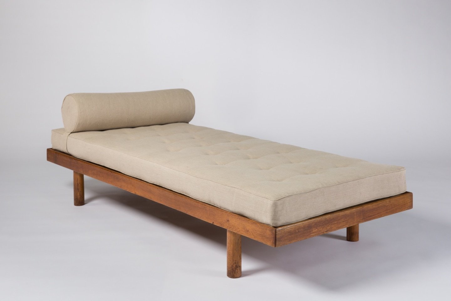Minimalist mid-century modern daybed with a beige tufted mattress and cylindrical bolster pillow, supported by a wooden frame and legs.