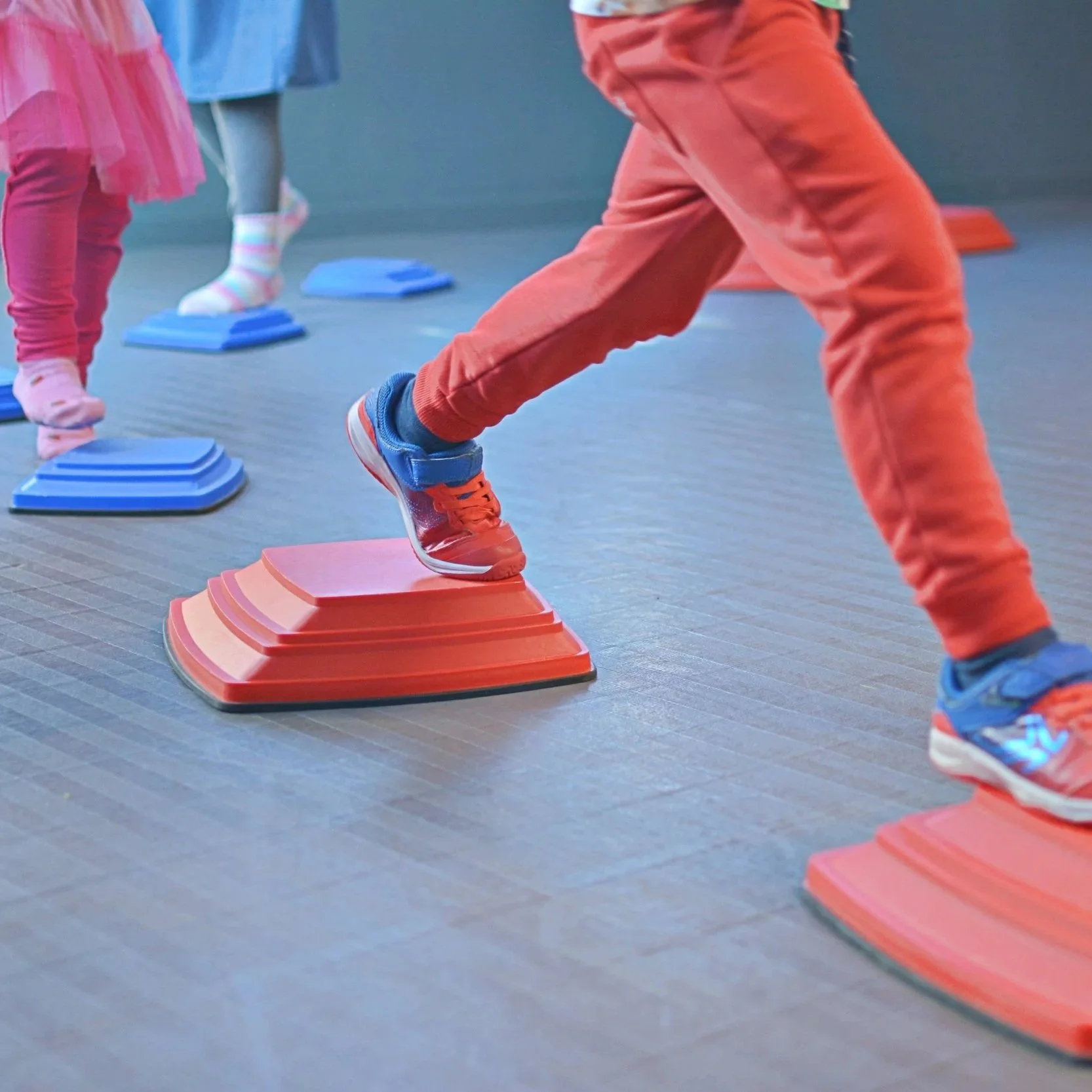 Children exercising on colorful aerobic step platforms in a gym, with a focus on the child's foot in sneakers stepping on a red platform.