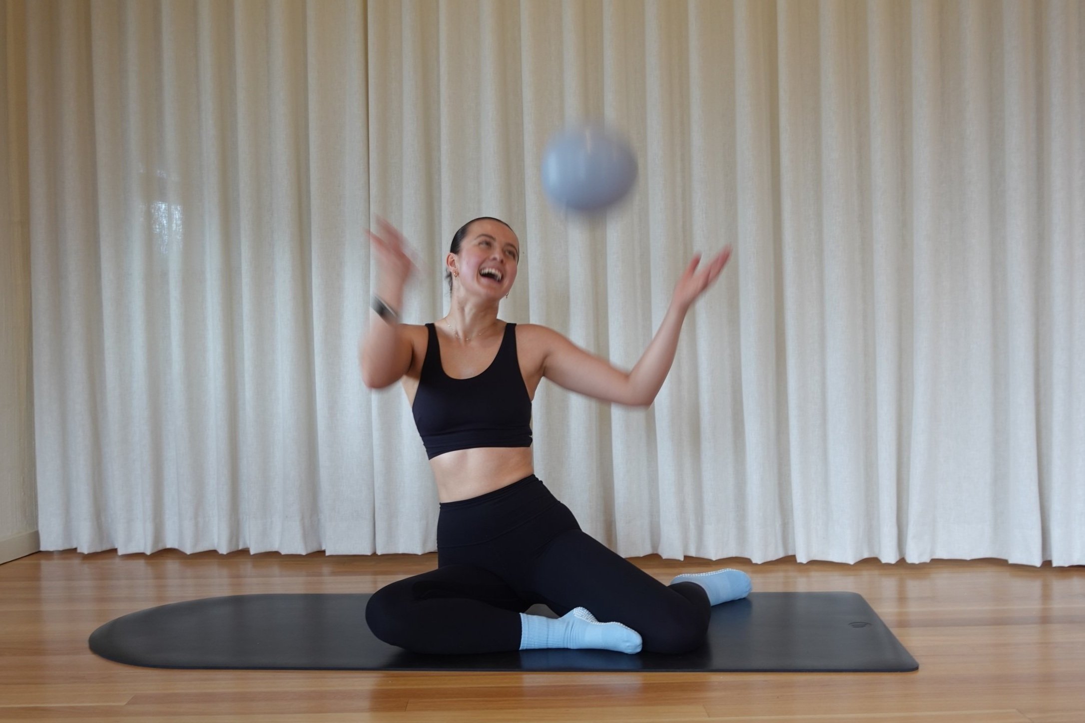 A woman sitting on a pilates mat in a black sports bra and leggings, smiling and playing with a light blue exercise ball in a room with beige curtains and wooden floors.