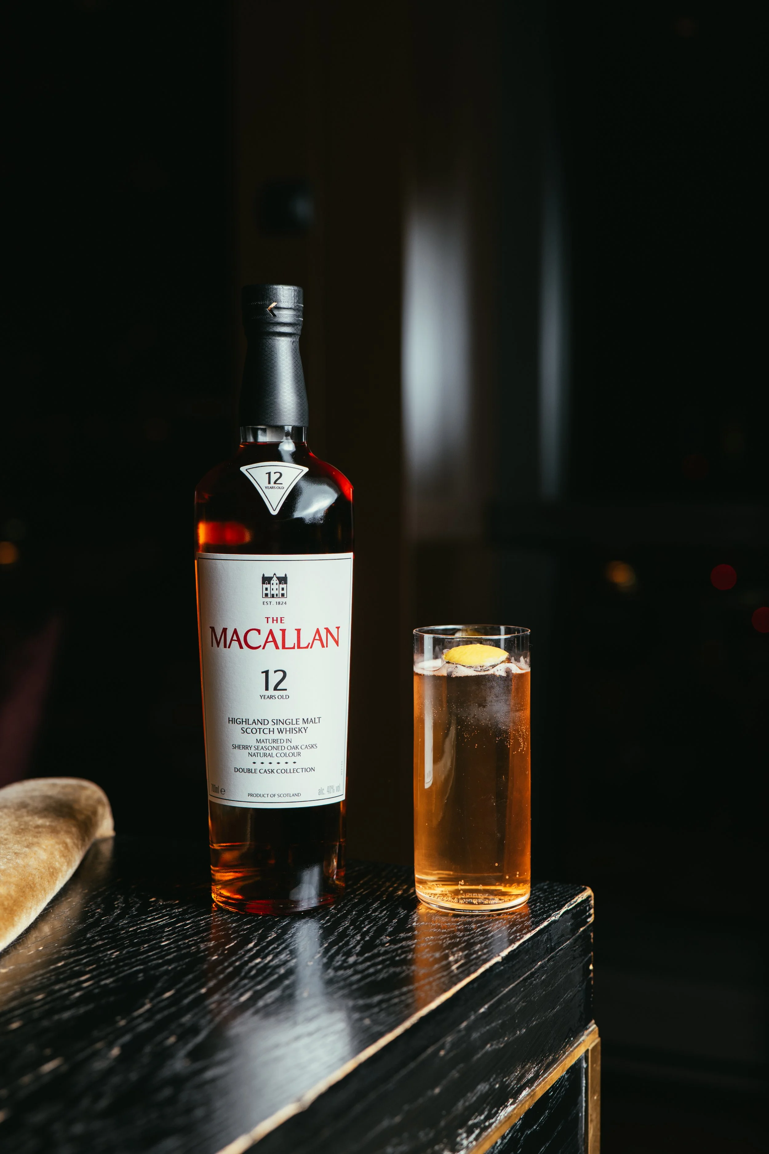 A bottle of The Macallan 12-year-old single malt scotch whisky next to a glass with a lemon garnish on a dark wooden surface.