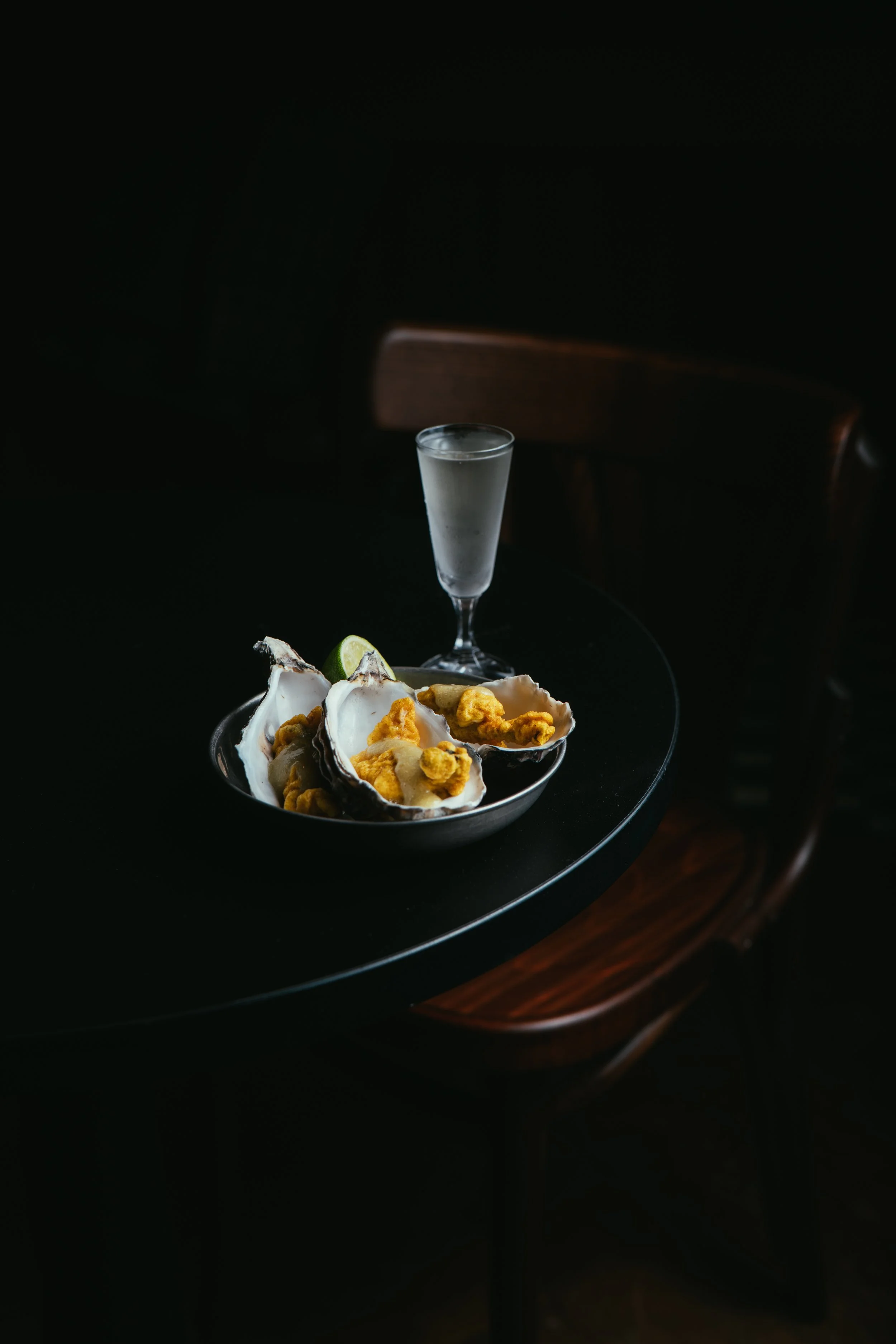 A dark table with a bowl of oysters topped with mignonette sauce and lemon wedges, and a glass of clear liquid, possibly water or a cocktail, in a dimly lit setting.
