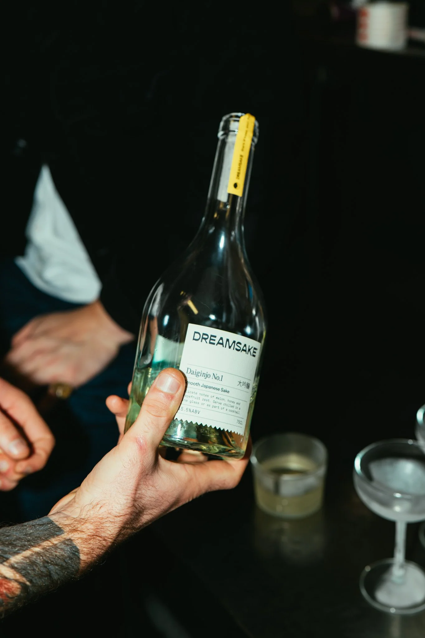 Person holding a bottle of Dreamake Daiginjo No. 1 Japanese sake in a dimly lit setting, with a glass of sake and a cocktail glass nearby.