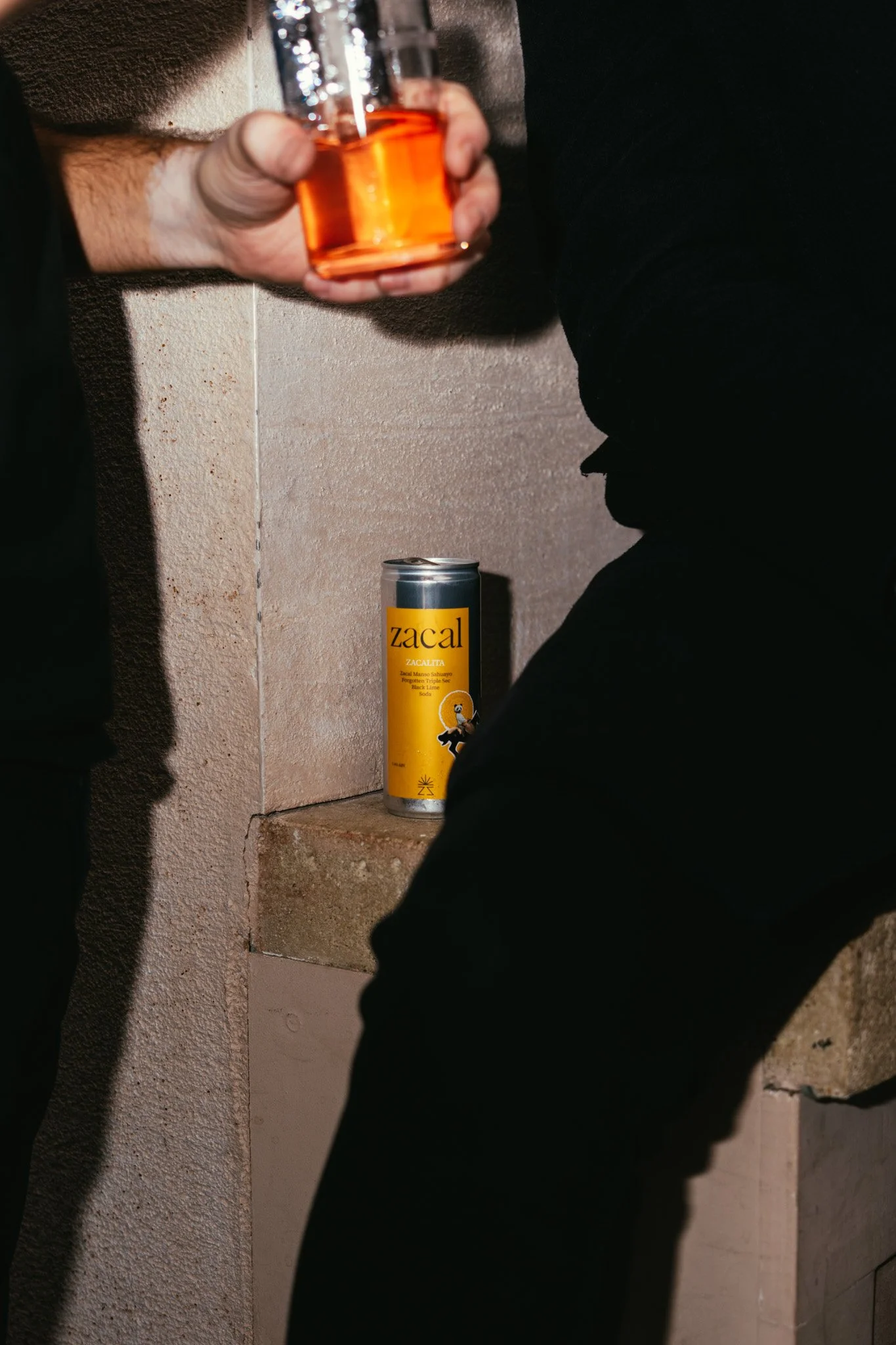 A hand holding a glass with an orange beverage, with a can of Zacal soda on a ledge in the background, set against a textured wall.