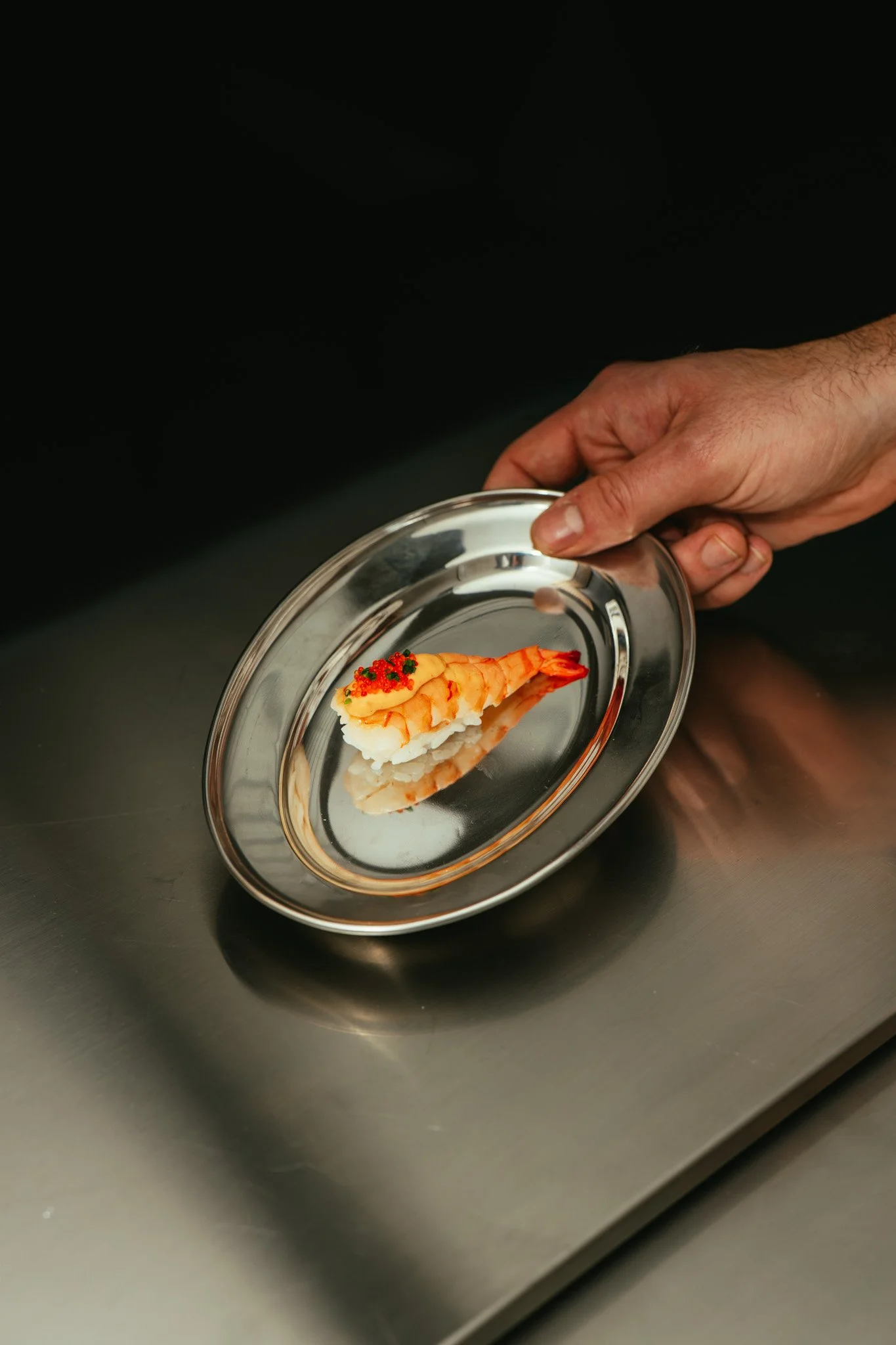 Close-up of a hand holding a small metal plate with a piece of sushi topped with shrimp and red fish roe.