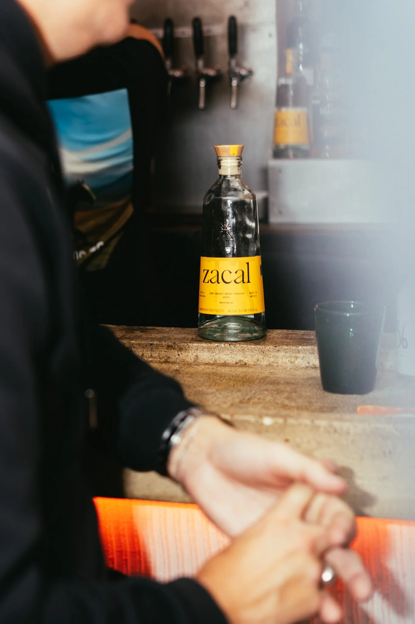 A glass bottle with a yellow label reading 'zacal' sitting on a wooden countertop with a black cup nearby, amidst a bar or kitchen setting, with a person sitting at the counter with their hands clasped.