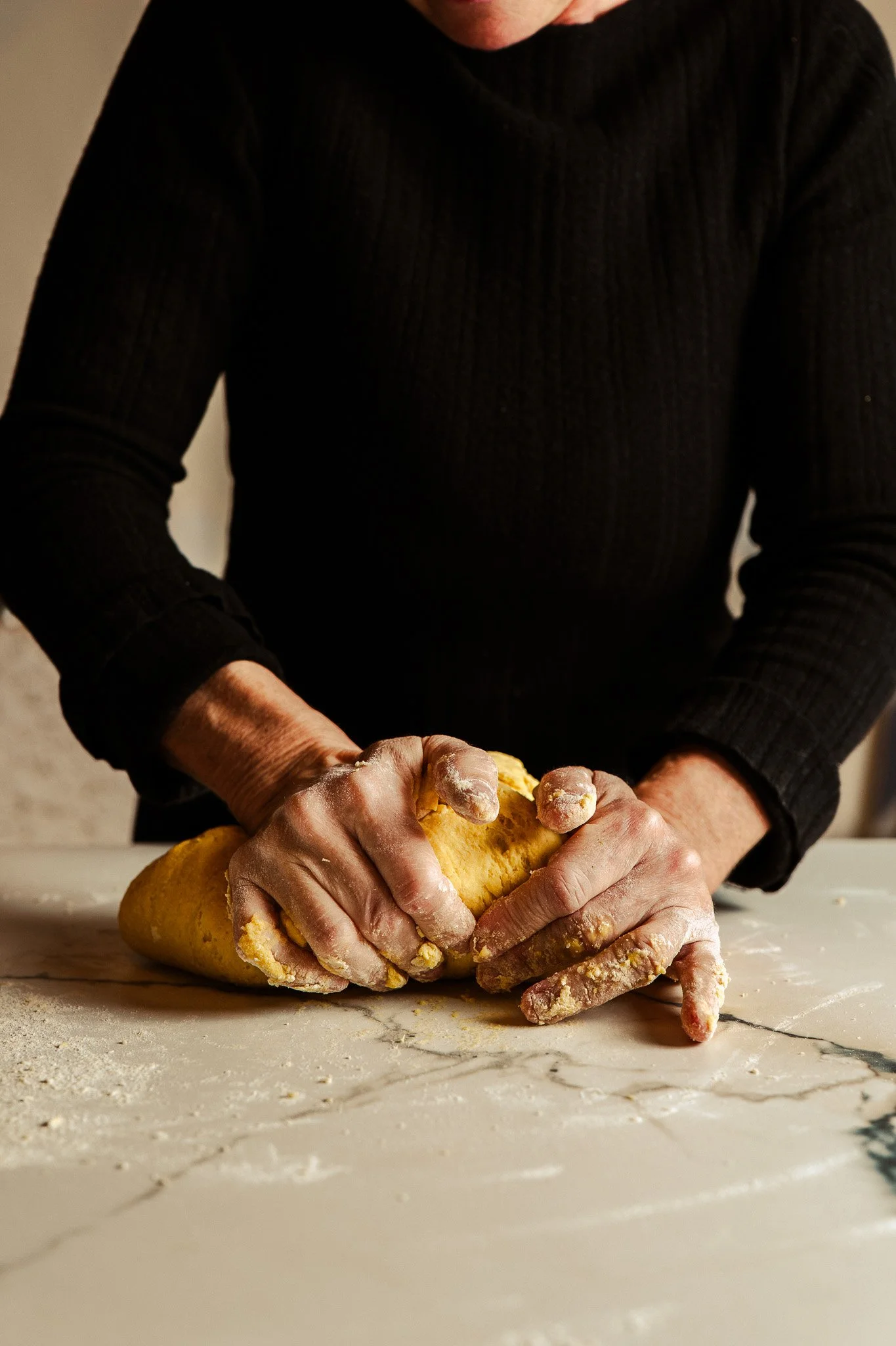 Person kneading dough on a marble countertop, wearing a black sweater, with flour on their hands.