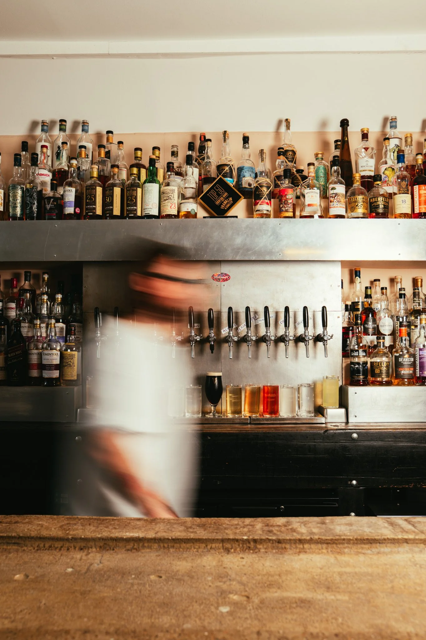 A blurred bartender leaning over a wooden bar counter with a row of colorful drinks behind the bar, and numerous bottles of liquor on shelves above.