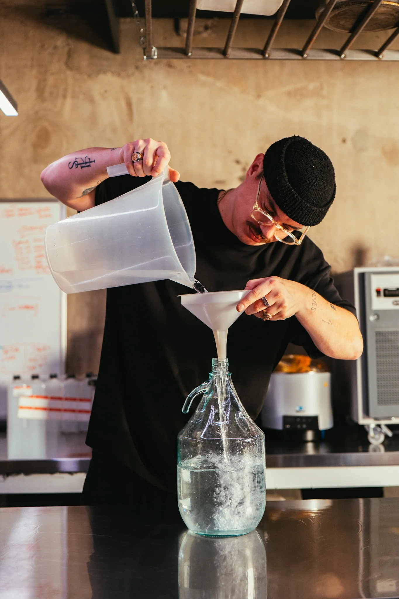 A man in black clothing, glasses, and a black beanie pouring a liquid from a clear jug through a funnel into a glass bottle, standing in a room with a beige wall and kitchen equipment.