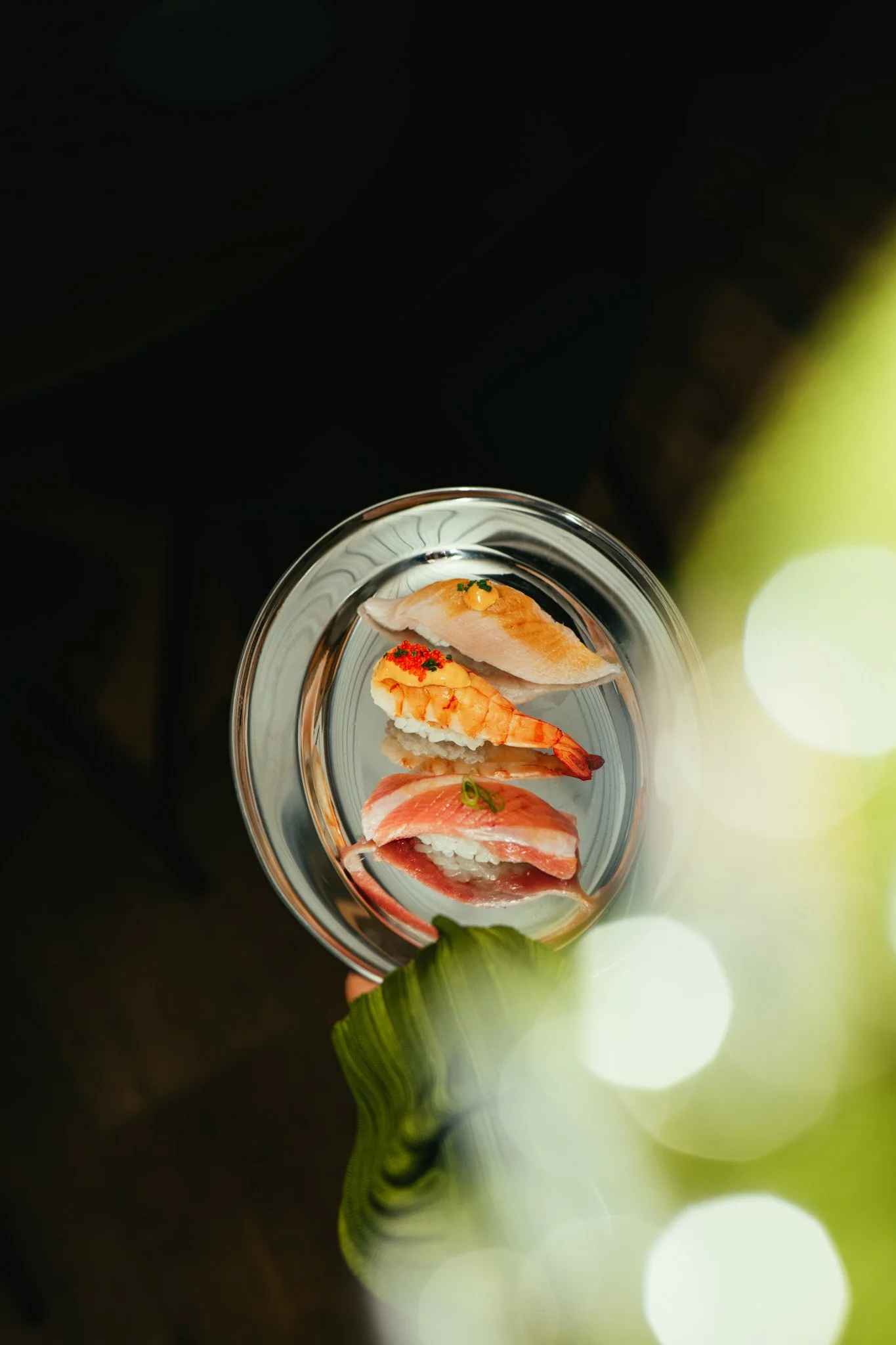 A glass plate with four different types of sushi arranged vertically, viewed from above with a blurred green leaf and bright bokeh effect in the foreground.
