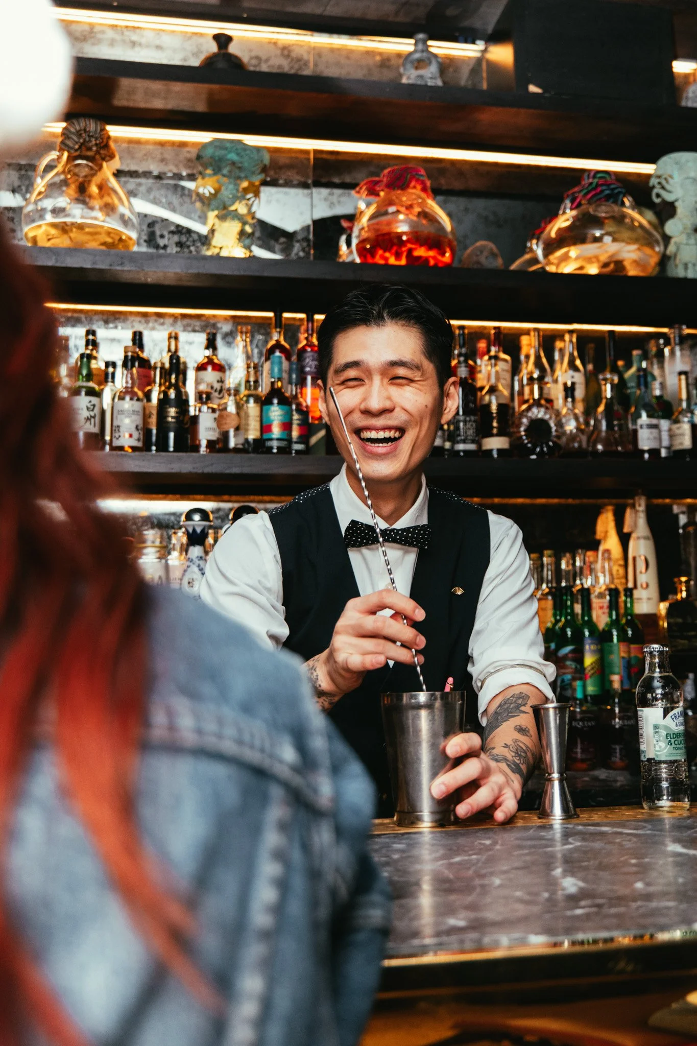 A bartender with tattoos and a bow tie smiling while working behind a bar, with bottles of liquor and bar tools in the background.