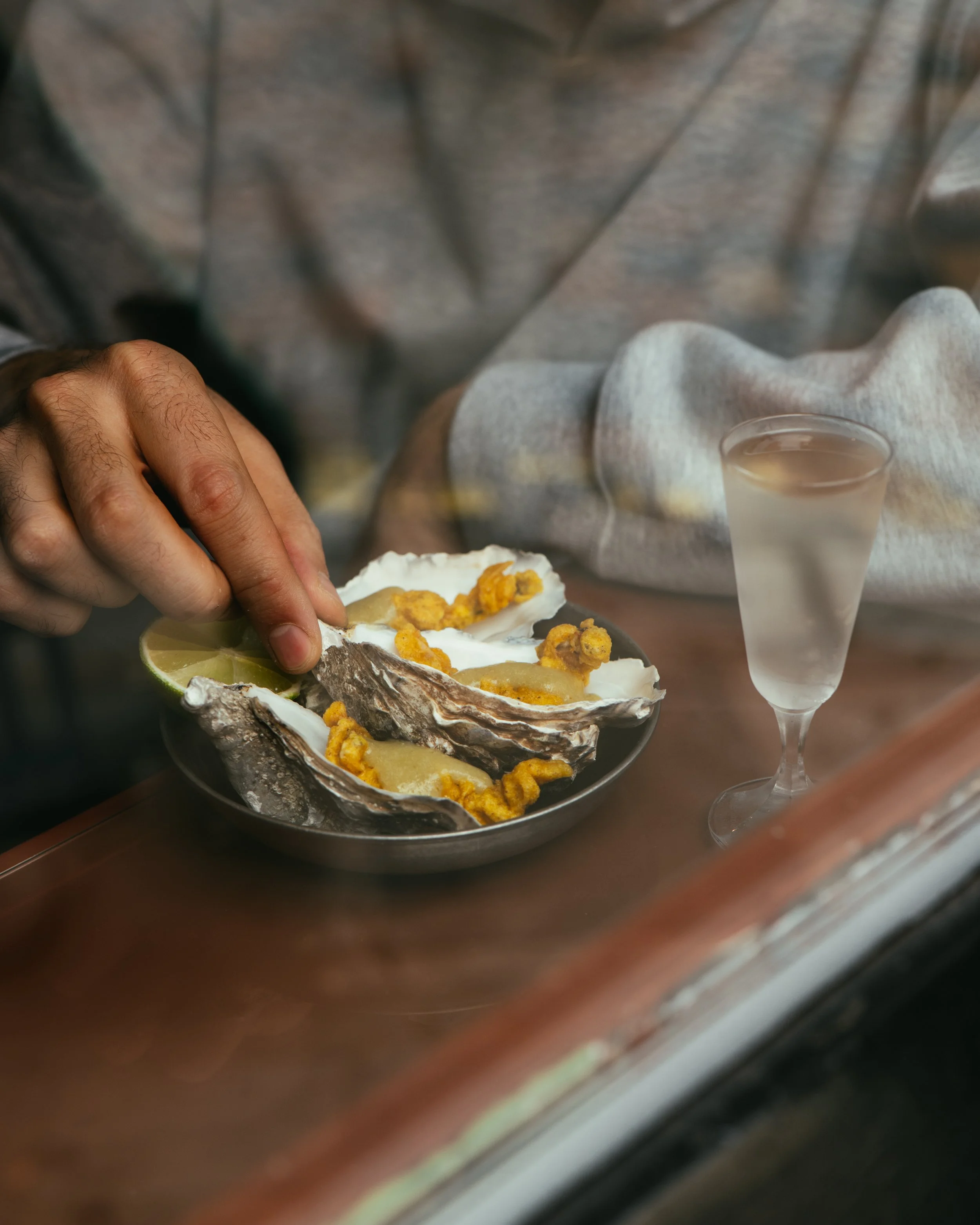 Person placing lemon wedge on oysters topped with fried garnishes, with a glass of clear beverage nearby.