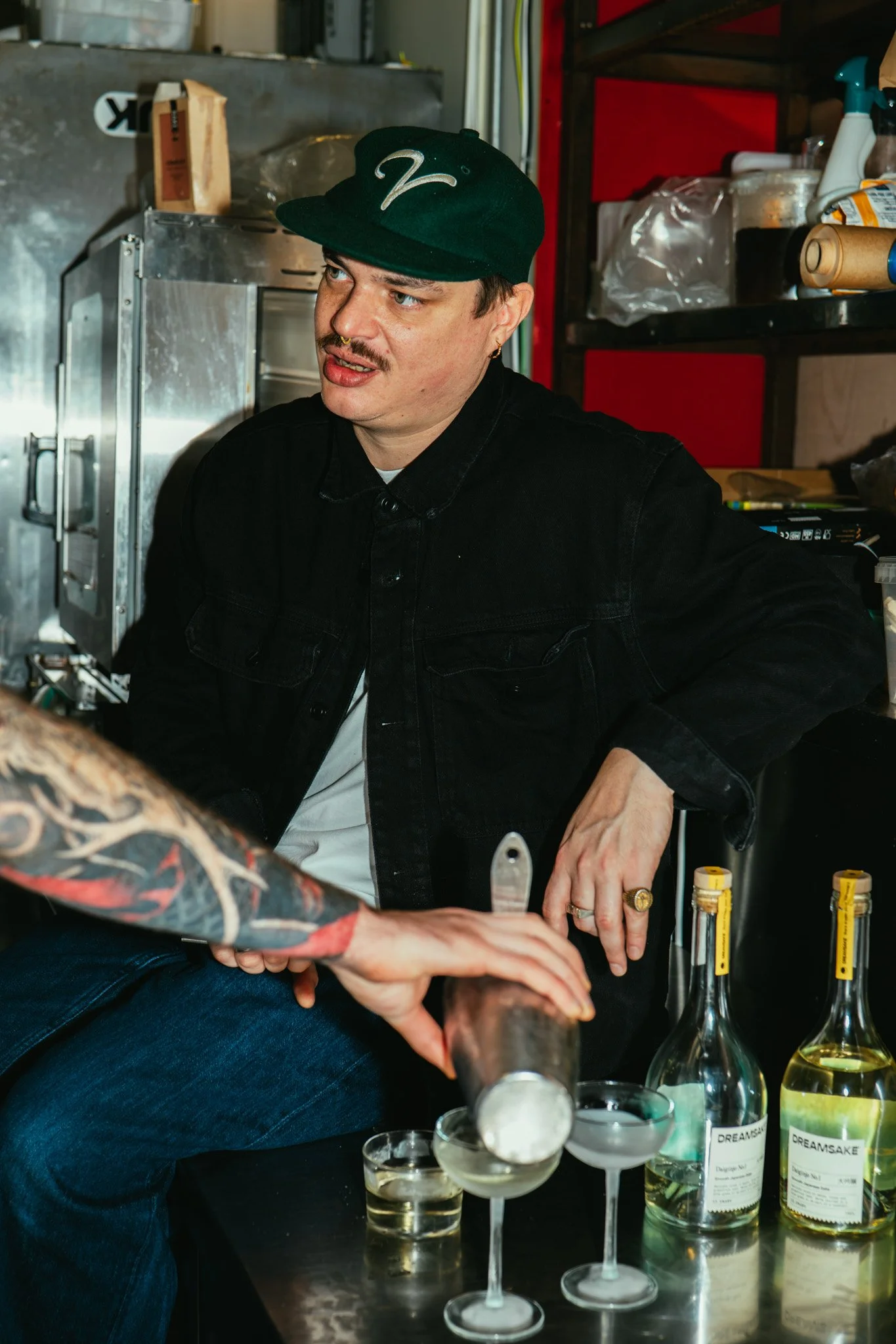 A man with a black hat, black jacket, and tattoos on his arm is sitting behind a bar, pouring a drink into cocktail glasses on a black table, with bottles labeled 'Dreamsake' nearby.