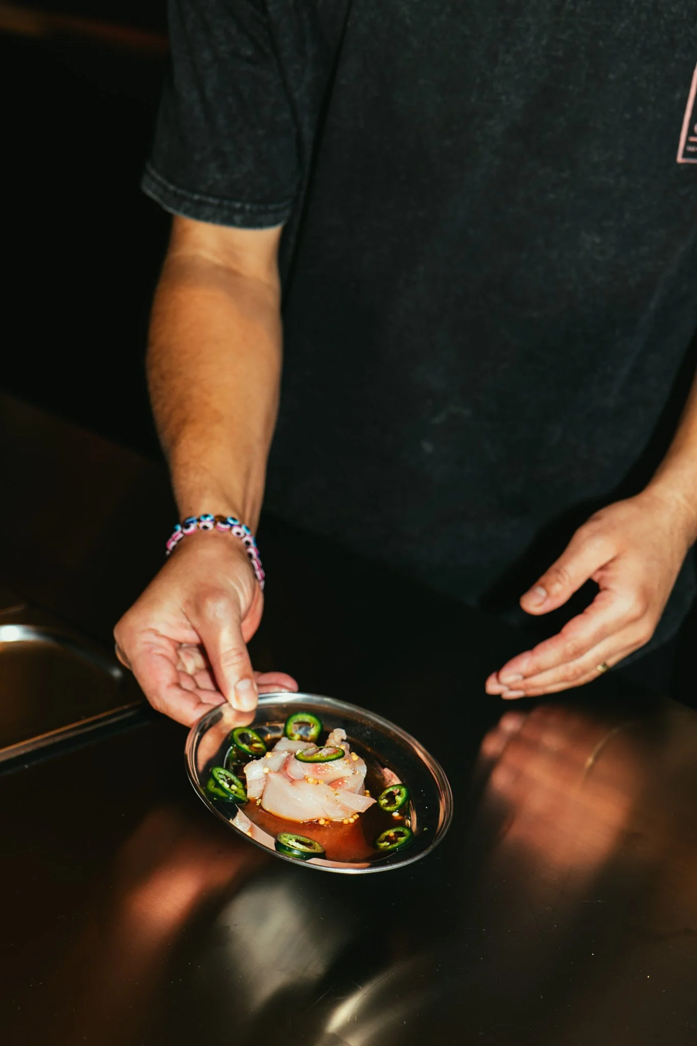 A person holding a small glass dish with raw fish, sliced jalapenos, and soy sauce.