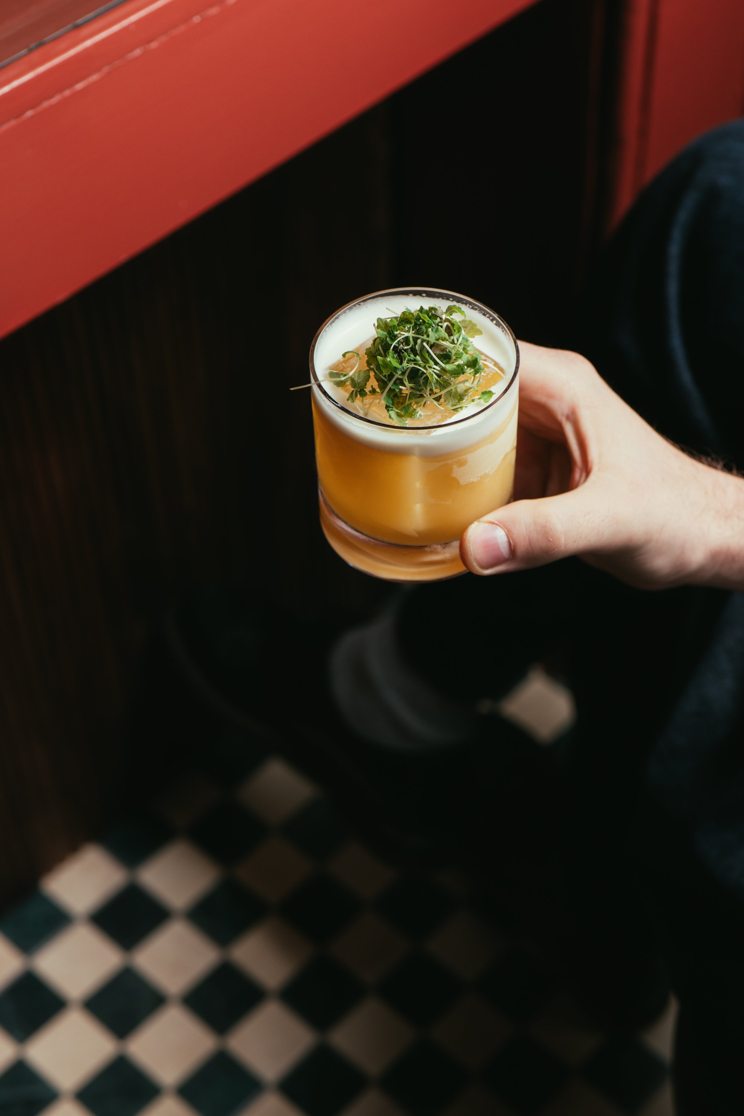 A hand holding a cocktail garnished with microgreens, against a background of a red wall and a black-and-white checkered floor.