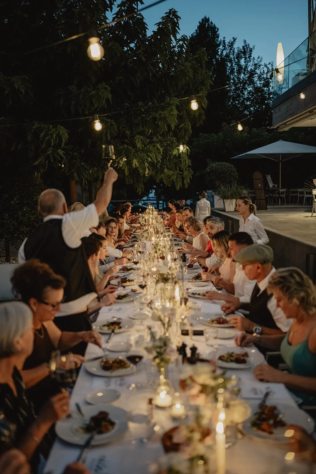 Eine lange gedeckte Tafel beim Abendessen im Freien, beleuchtet mit Lichterketten, mit vielen Gästen und einem Kellner, der einen Drink serviert.