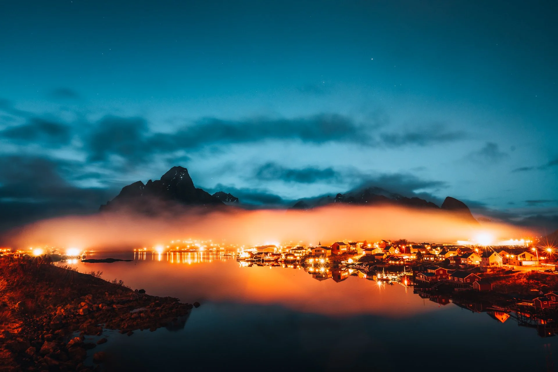 Ein nächtliches Bild eines kleinen Hafens mit beleuchteten Häusern, Bergen im Hintergrund und einem bewölkten Himmel.