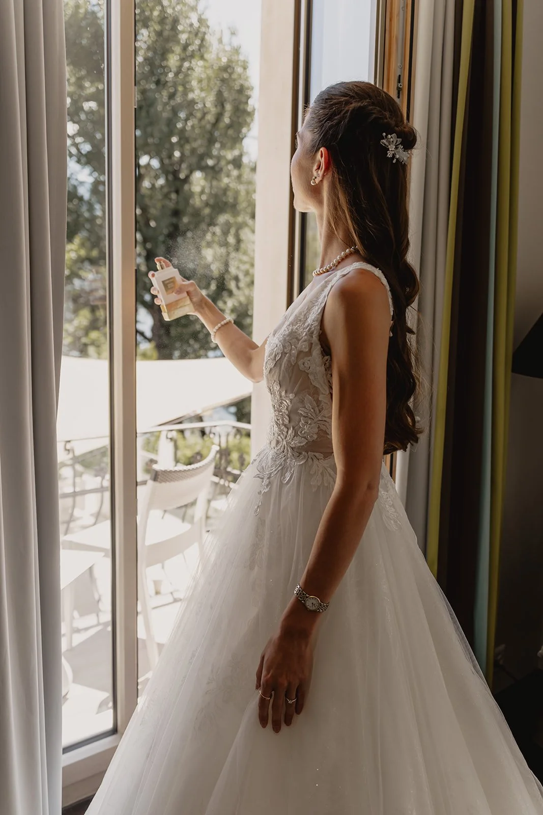 Eine Frau in einem weißen Brautkleid sprüht Parfüm auf sich, während sie vor einem Fenster steht. Sie trägt Perlenschmuck, eine Uhr und hat lange, braune Haare.