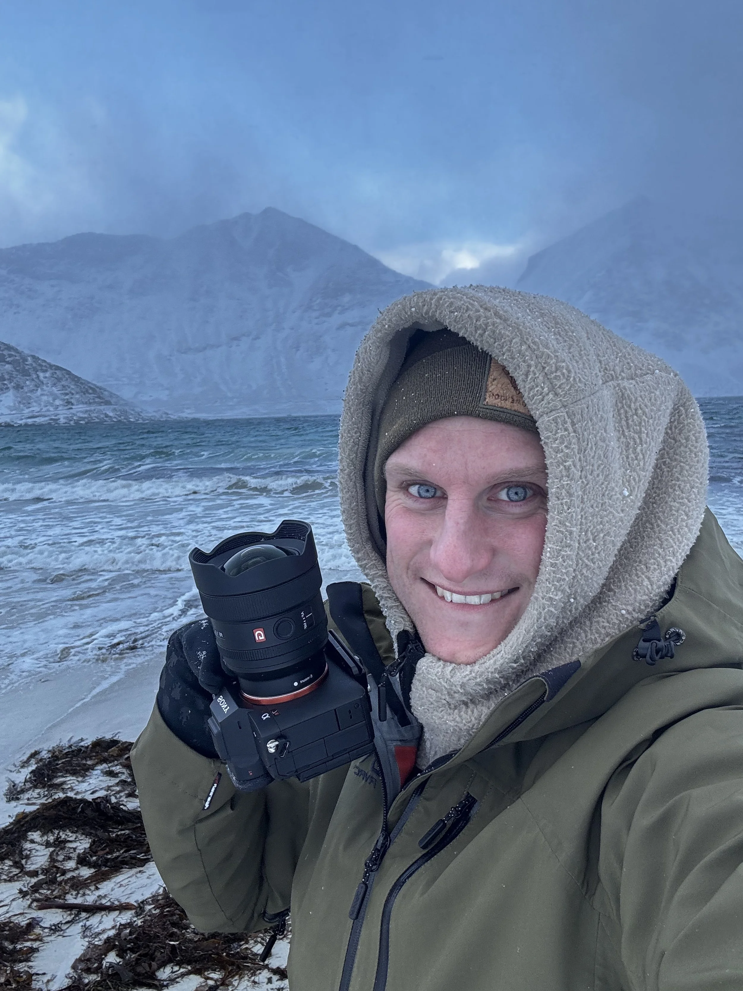 Ein Mann im Winterkleidung, der an einem felsigen Strand mit Wasser und Bergen im Hintergrund fotografiert.