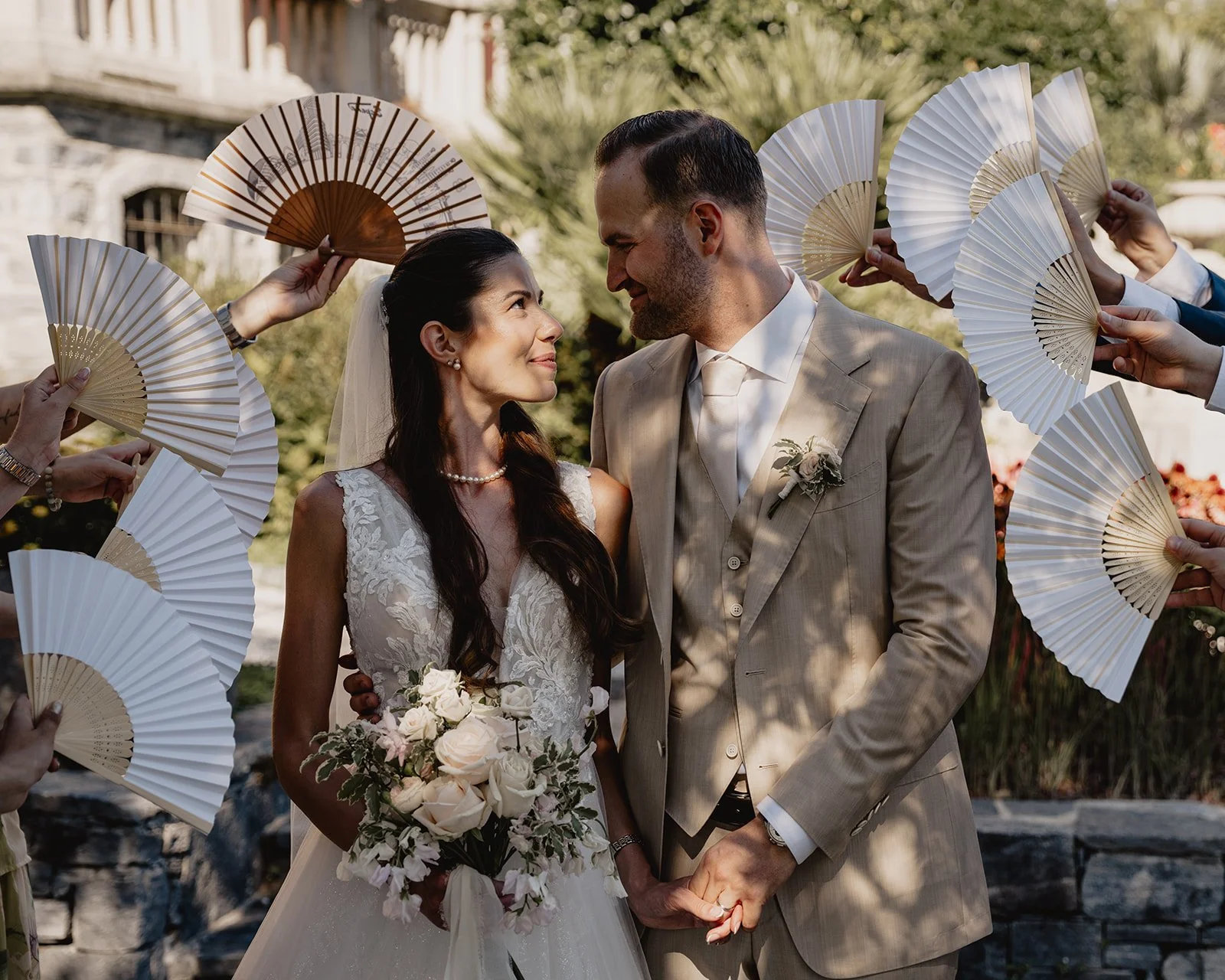 Ein Heiratspaar hält sich bei ihrer Hochzeit die Hände und blickt sich liebevoll an, umgeben von schwenkenden Fächern.