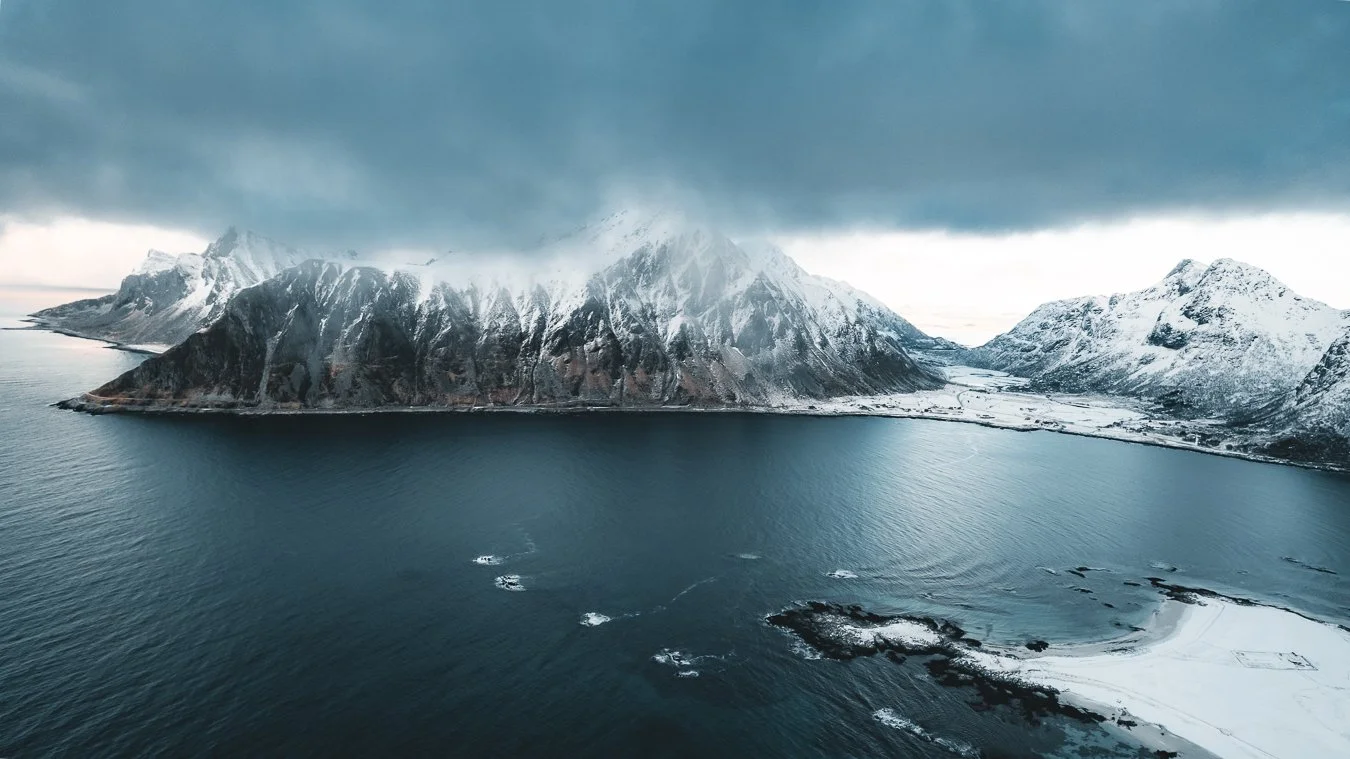 Berglandschaft mit schneebedeckten Gipfeln, Wasser im Vordergrund und dunklem Himmel