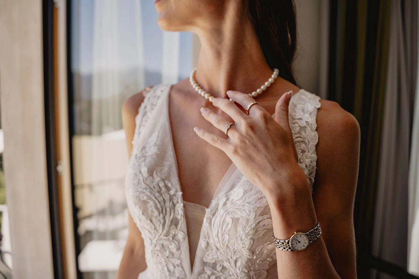 Frau in weißem Spitzenkleid trägt Perlenkette, Ringe und Armbanduhr, berührt das Dekolleté, im Hintergrund Fenster mit Blick nach draußen.