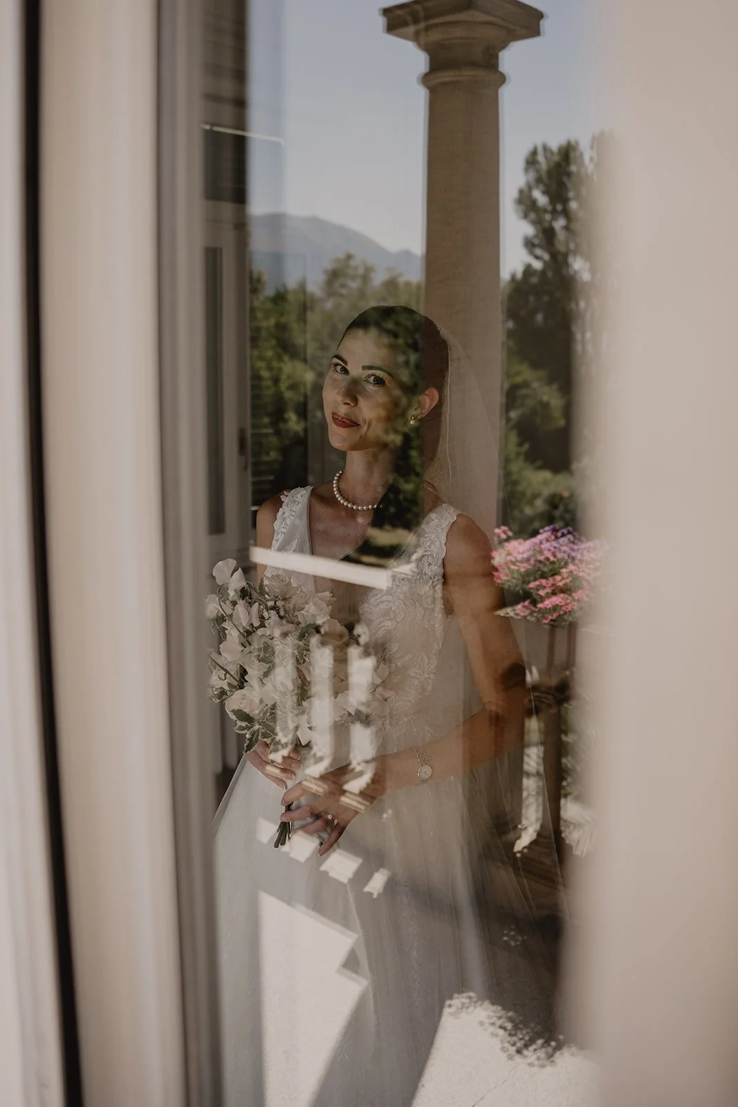 Braut in Hochzeitskleid schaut durch eine Fensterscheibe, hält einen Blumenstrauß, mit einer Landschaft im Hintergrund.