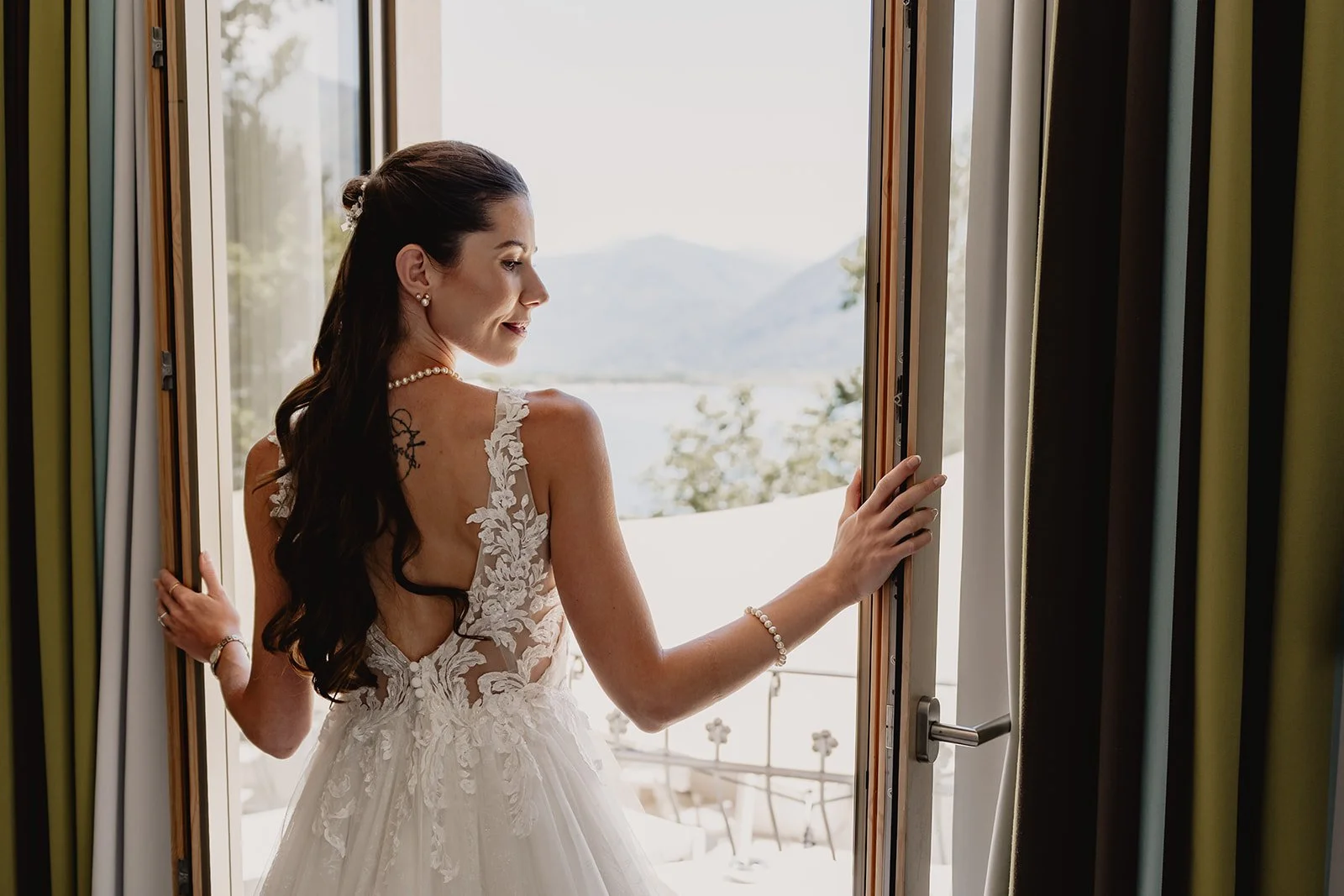 Eine Braut in einem weißen Hochzeitskleid steht an einem offenen Fenster, blickt nach draußen auf eine Landschaft mit Wasser und Bergen, während sie die Fensterläden hält.