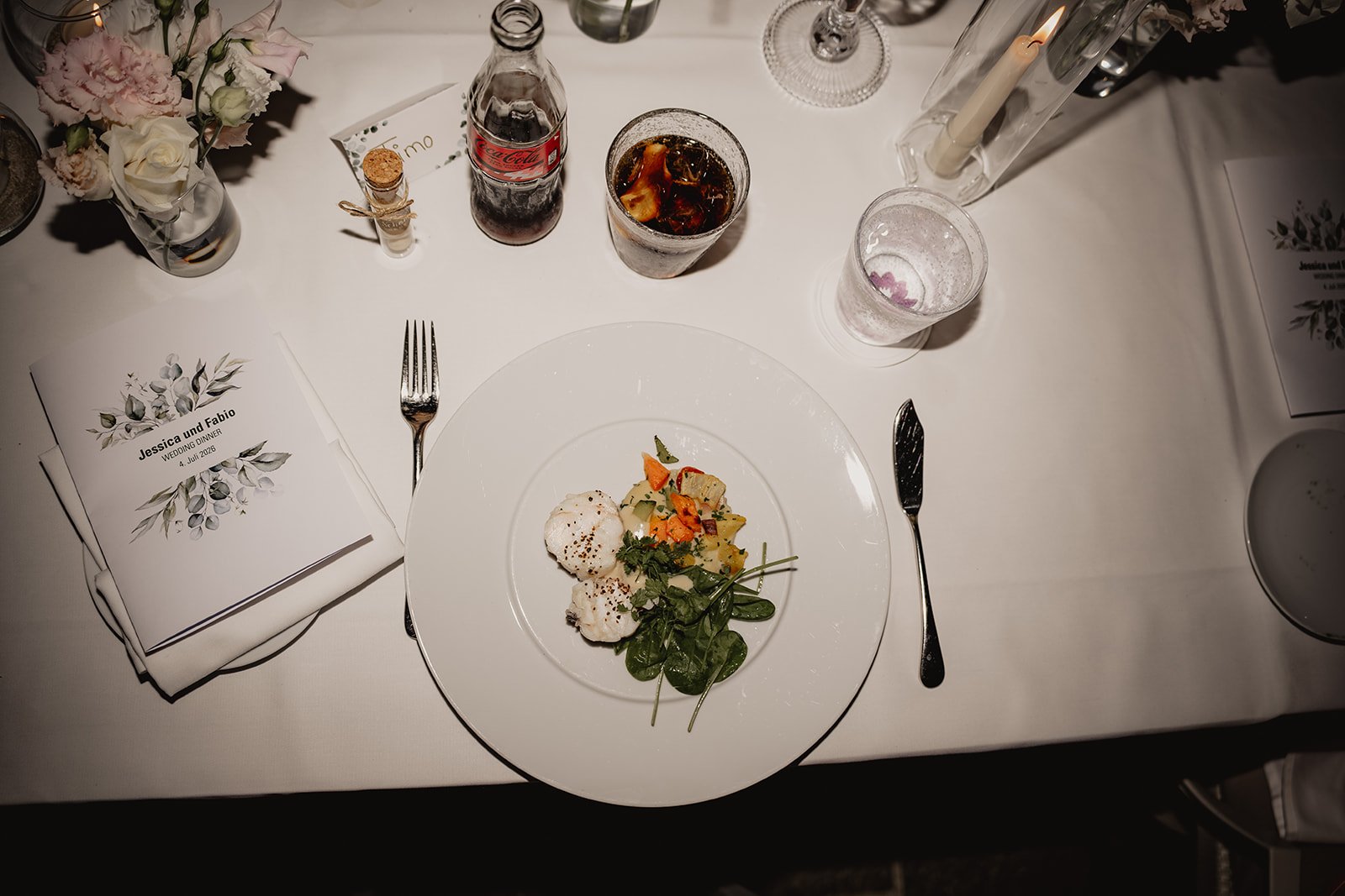 Ein festlich gedeckter Tisches mit einem weißen Teller, auf dem eine Portion Essen, bestehend aus einem Salat mit verschiedenem Gemüse und zwei Bällchen, serviert wird. Es gibt Besteck an beiden Seiten des Tellers, eine Brosche mit Blumen und eine Ma