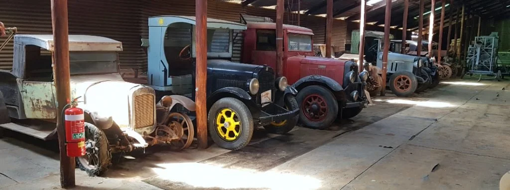Wyalkatchem CBH Museum wheatbin, Old, vintage cars stored in a barn with wooden support beams and a concrete floor.
