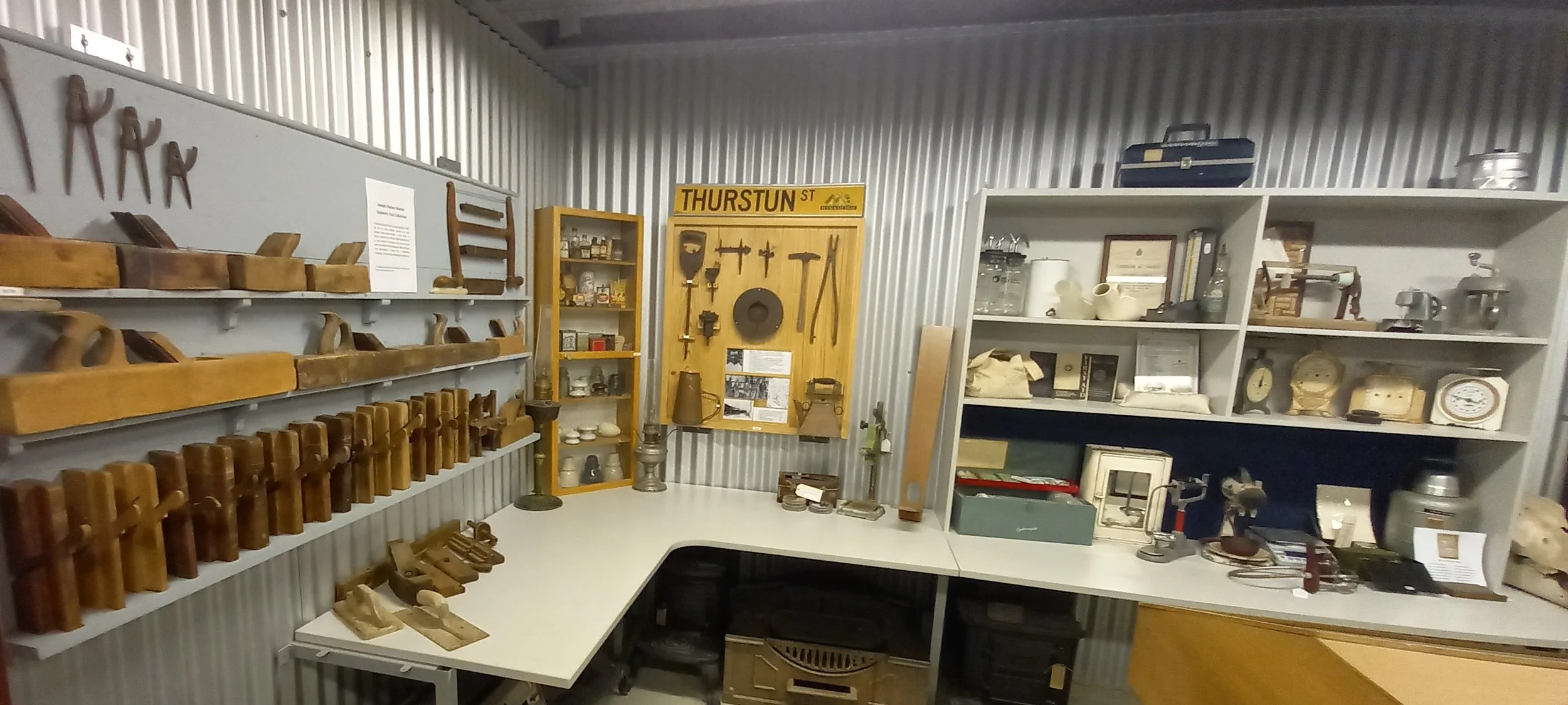 Wyalkatchem CBH Museum. A room with tools and objects on shelves, including woodworking tools like planes, a yellow display with vintage tools, and various framed and unframed items on the shelves.