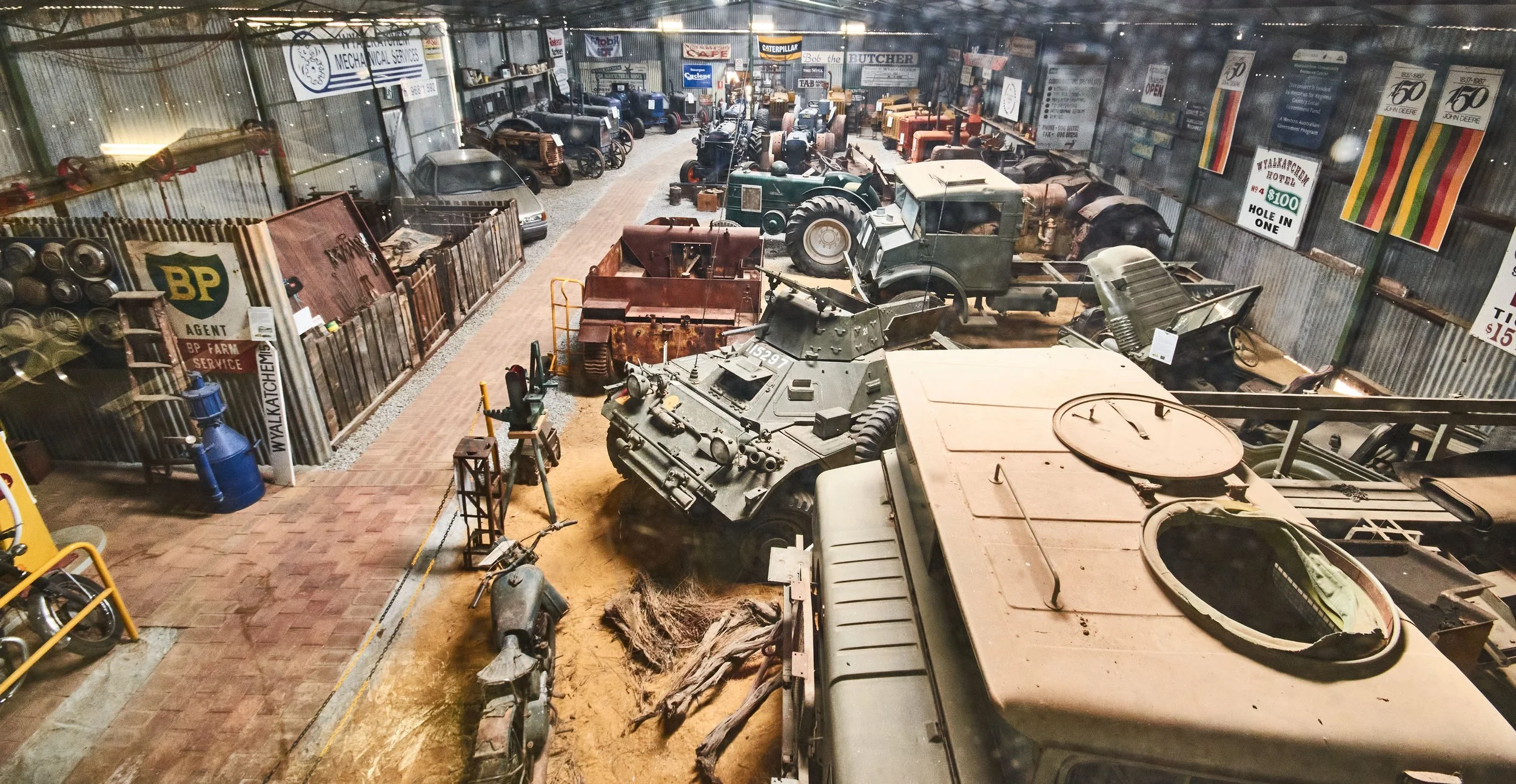 Vern's shed, Indoor vintage vehicle and machinery museum with various old trucks, tractors, and car parts displayed along a brick pathway.