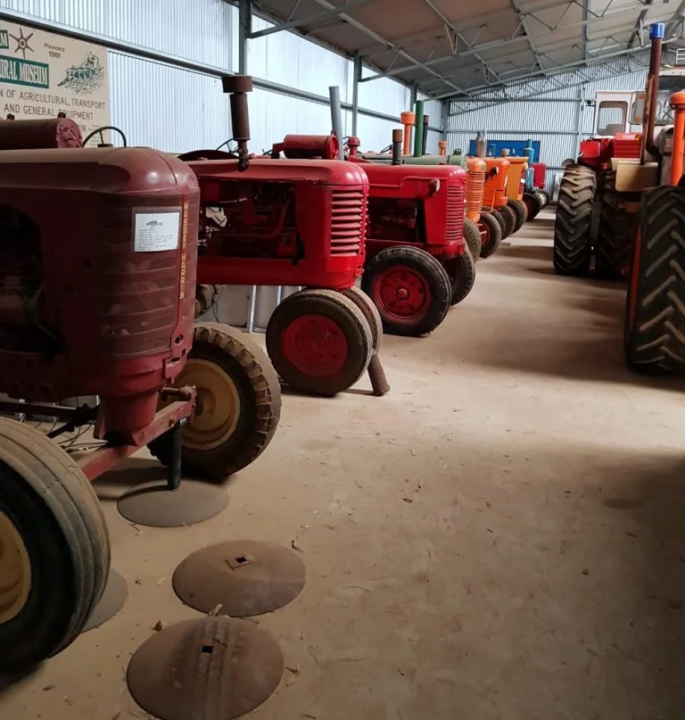 Kevin's shed, Wyalkatchem CBH Museum. A lineup of vintage tractors, mainly red, inside a large barn or shed with a high ceiling and metal walls.