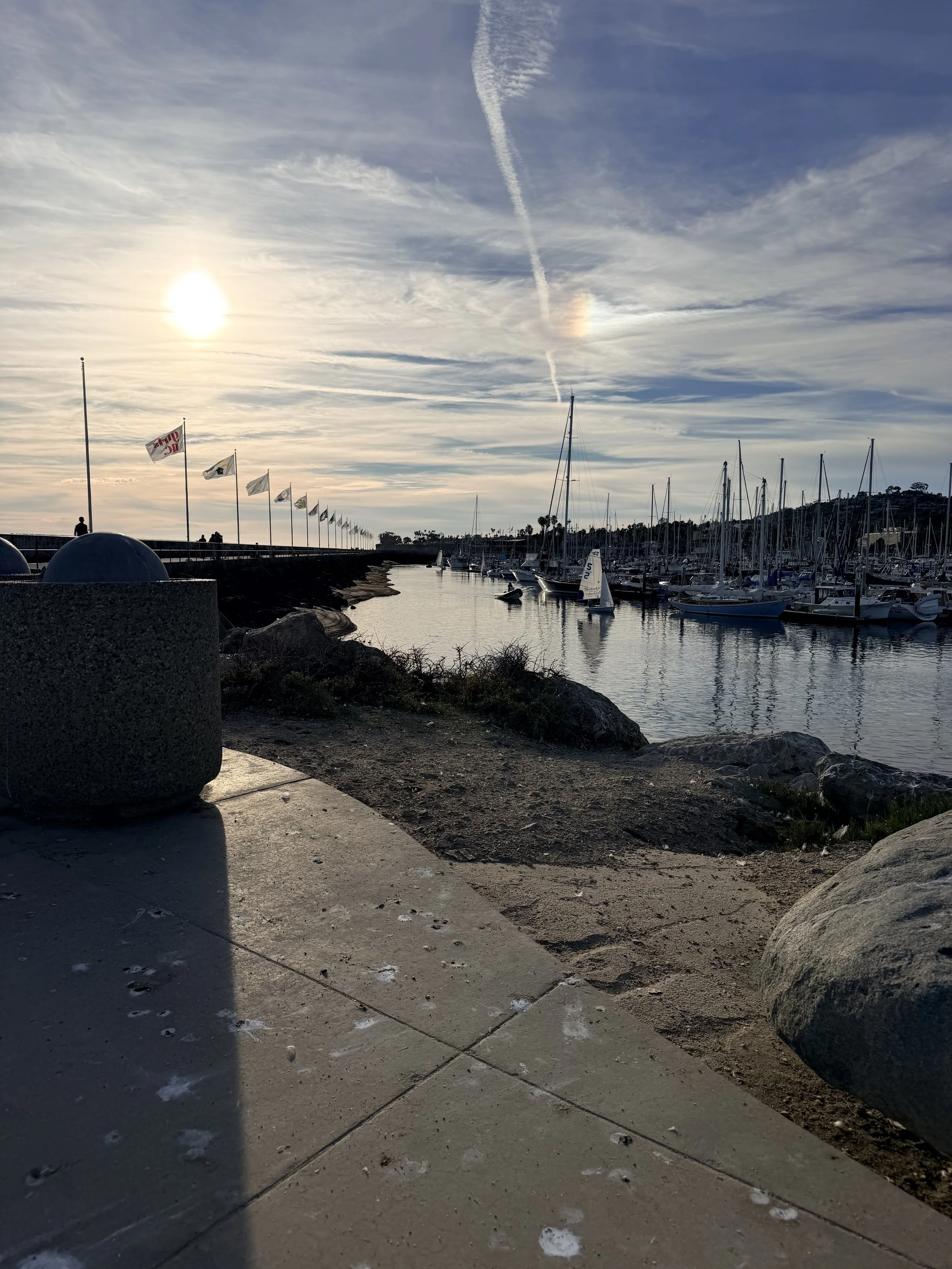 Sunset view of a marina with sailboats and yachts docked, flags along the pier, a lighthouse in the distance, and clouds in the sky.