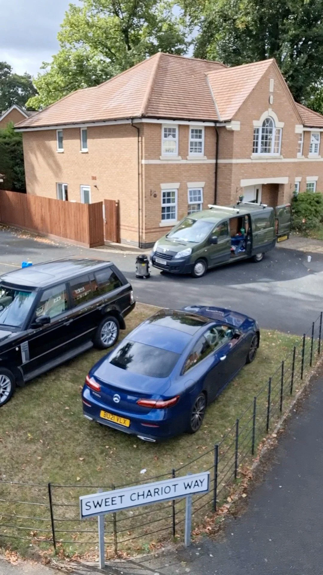 A residential street with a brick house and four parked cars, including a black estate, a blue sedan, and an open green delivery van, with a street sign saying 'Sweet Chariot Way'. Clean Roof.