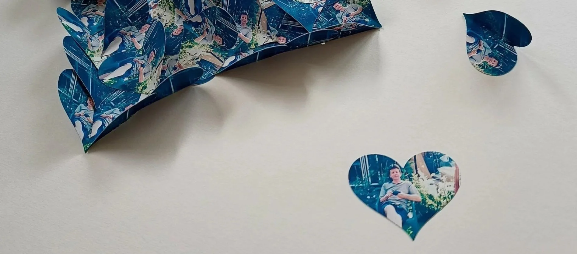 A partially assembled 3D paper heart with a replicated photo pattern, along with cut-out paper pieces including a heart-shaped piece displaying a man sitting outdoors with trees and plants in the background.