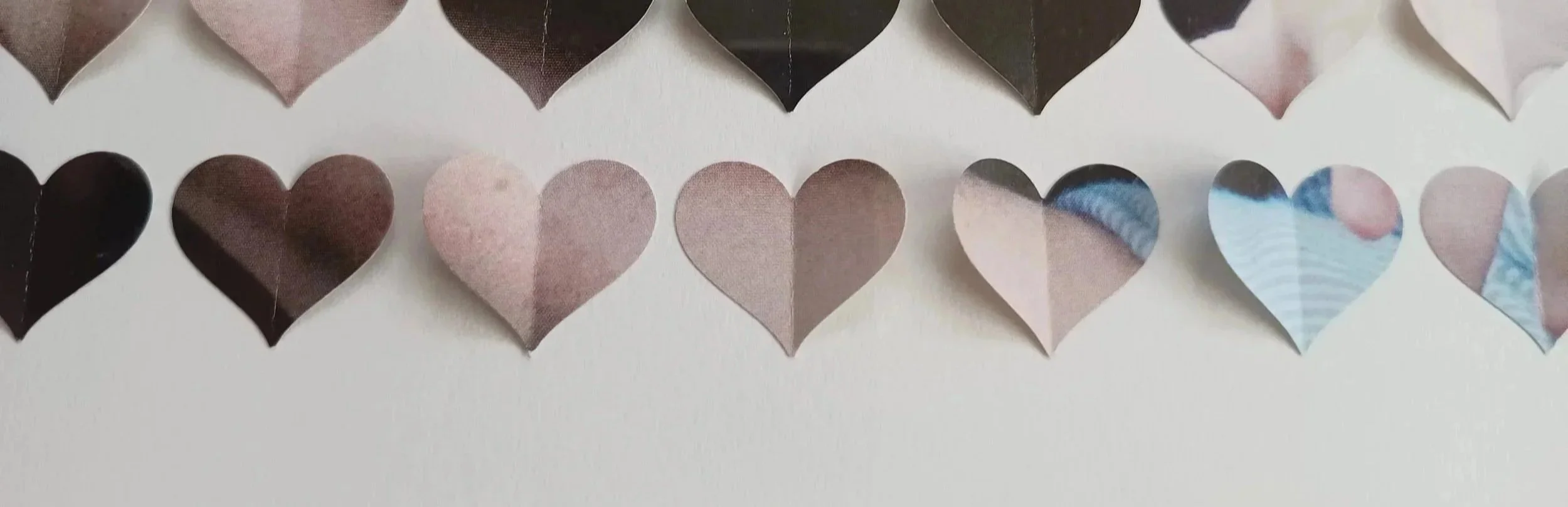 A row of paper hearts in various shades of black, brown, pink, and blue, arranged on a white surface.