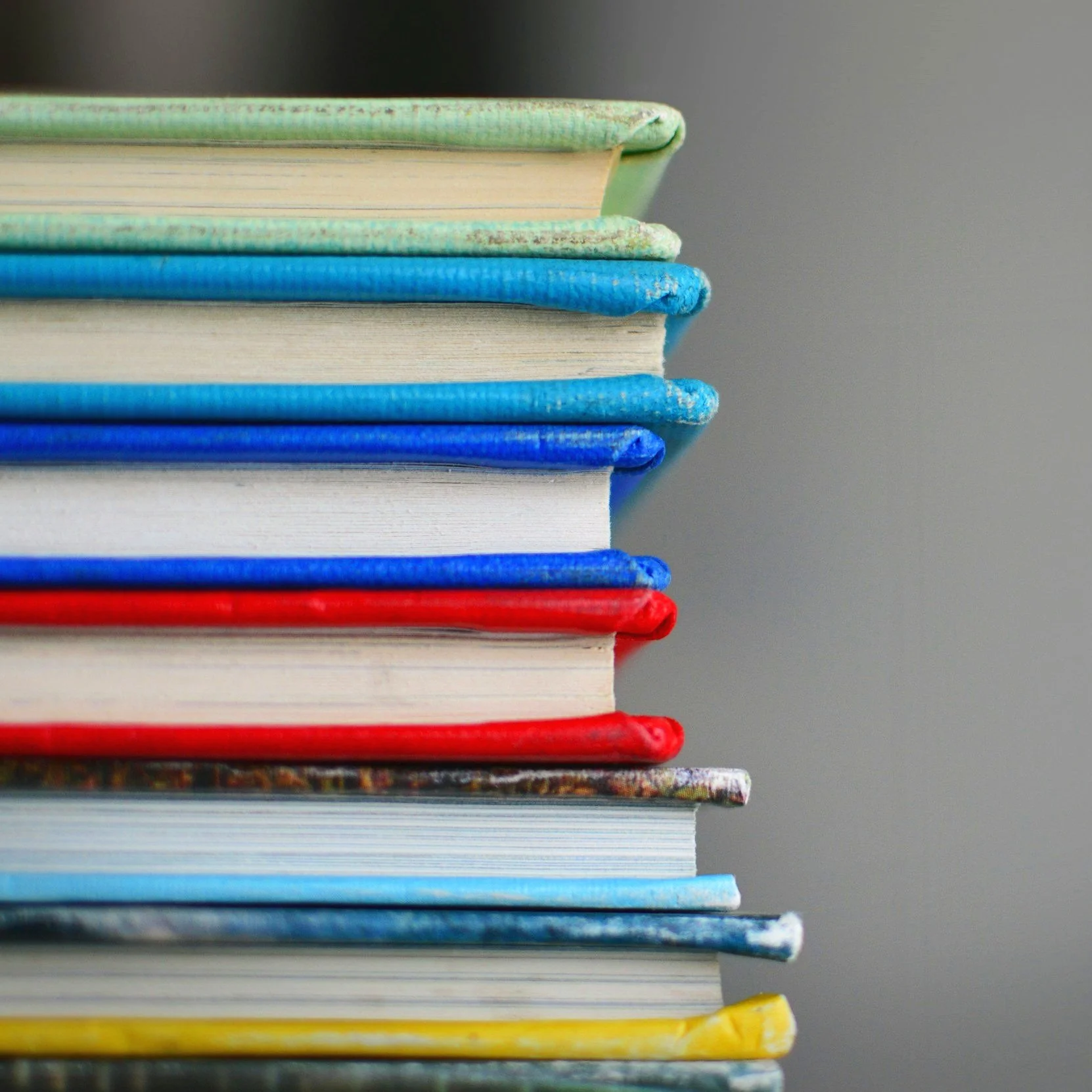 Stack of colorful books with covers in various colors including green, blue, white, red, gray, and yellow, stacked vertically against a gray background.