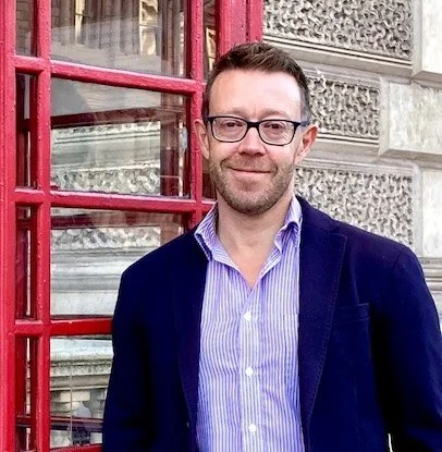 An expert mathematics coach. He is standing outside near red telephone booth, wearing a purple shirt and navy blazer.