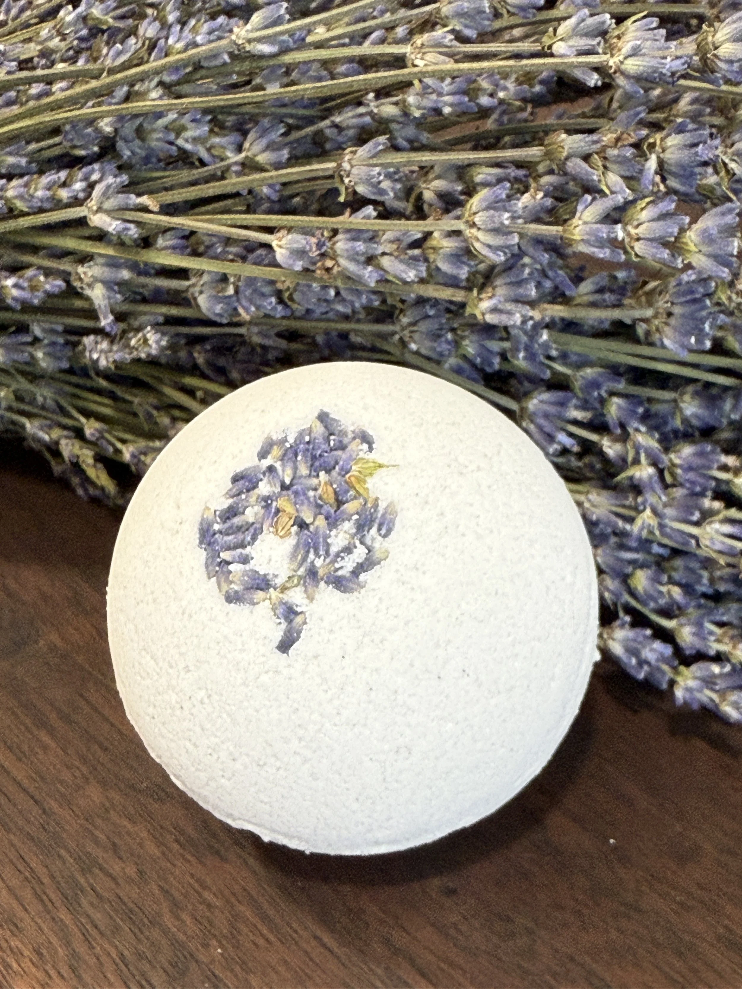 A white bath bomb with dried lavender flower decoration on top, placed on a wooden surface with a bunch of dried lavender flowers in the background.