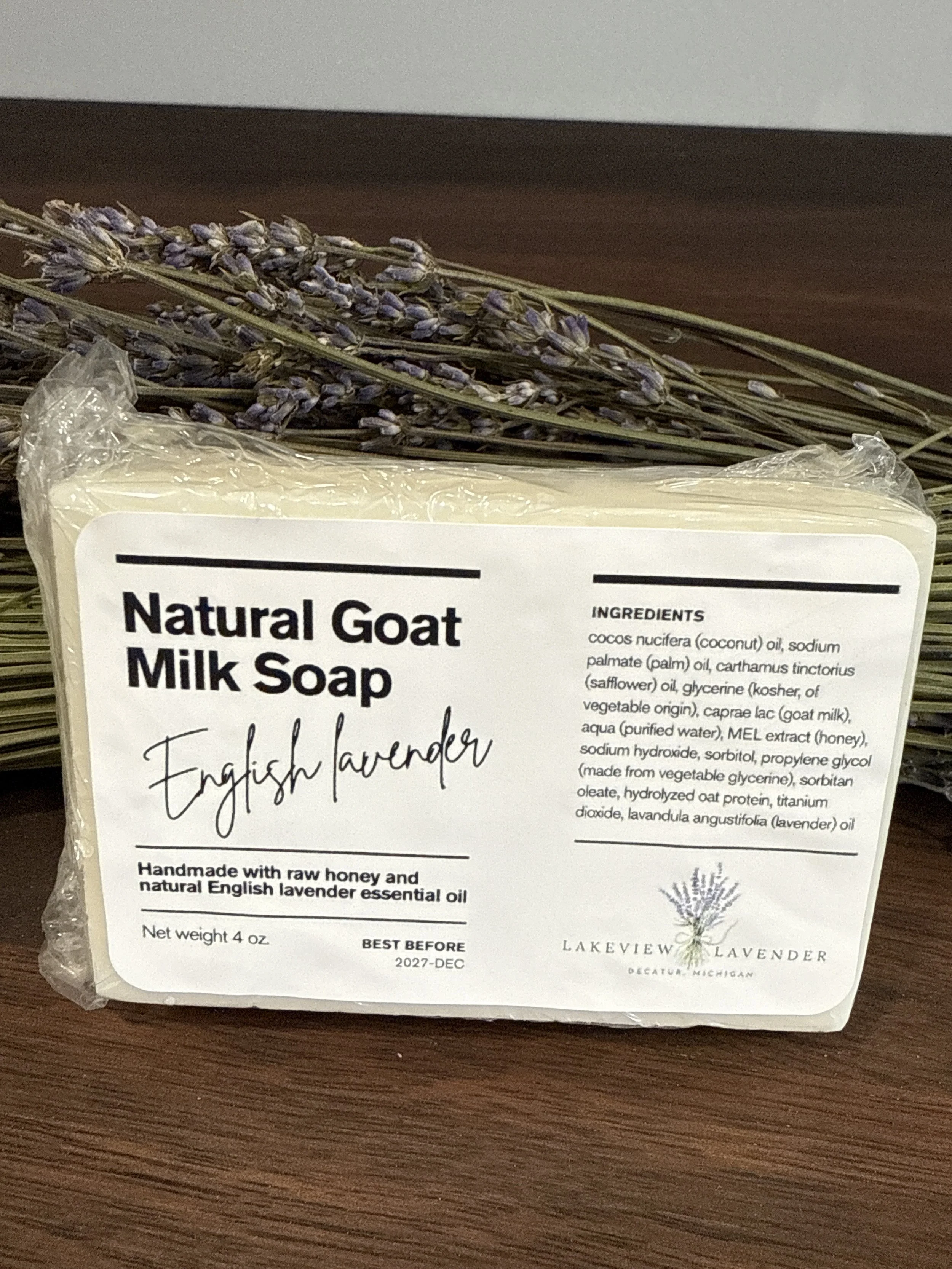 A bar of Natural Goat Milk Soap in front of a bundle of dried lavender flowers. The soap is labeled as English Lavender scent with ingredients listed, and placed on a wooden surface.