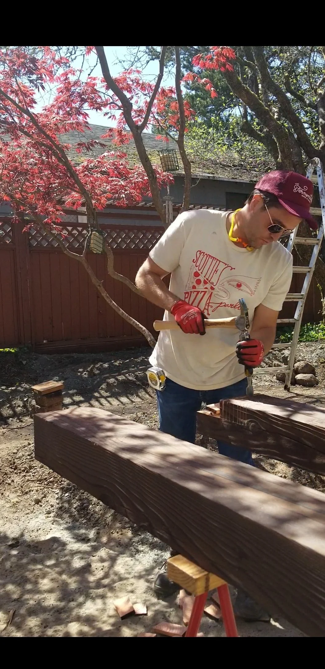 A person with sunglasses, a maroon cap, and a white T-shirt using a hammer and chisel to carve wood outdoors. They are wearing red work gloves and a yellow safety necklace. There is a ladder and a wooden fence in the background, along with trees with red and green leaves.