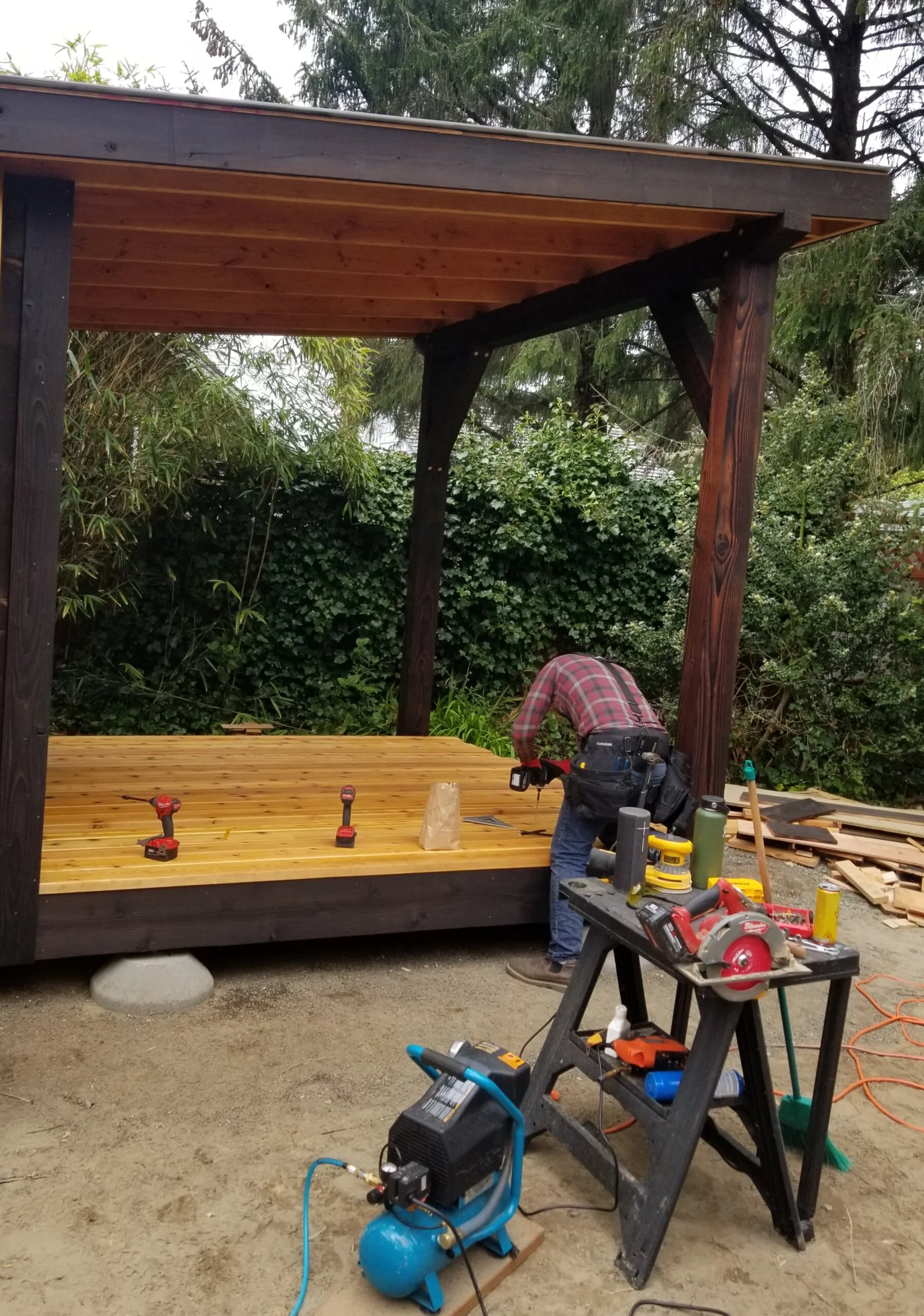 A person is installing a wooden deck under a partially built outdoor structure with a wooden roof and Shou Sugi Ban dark-stained support beams, surrounded by green bushes and trees.